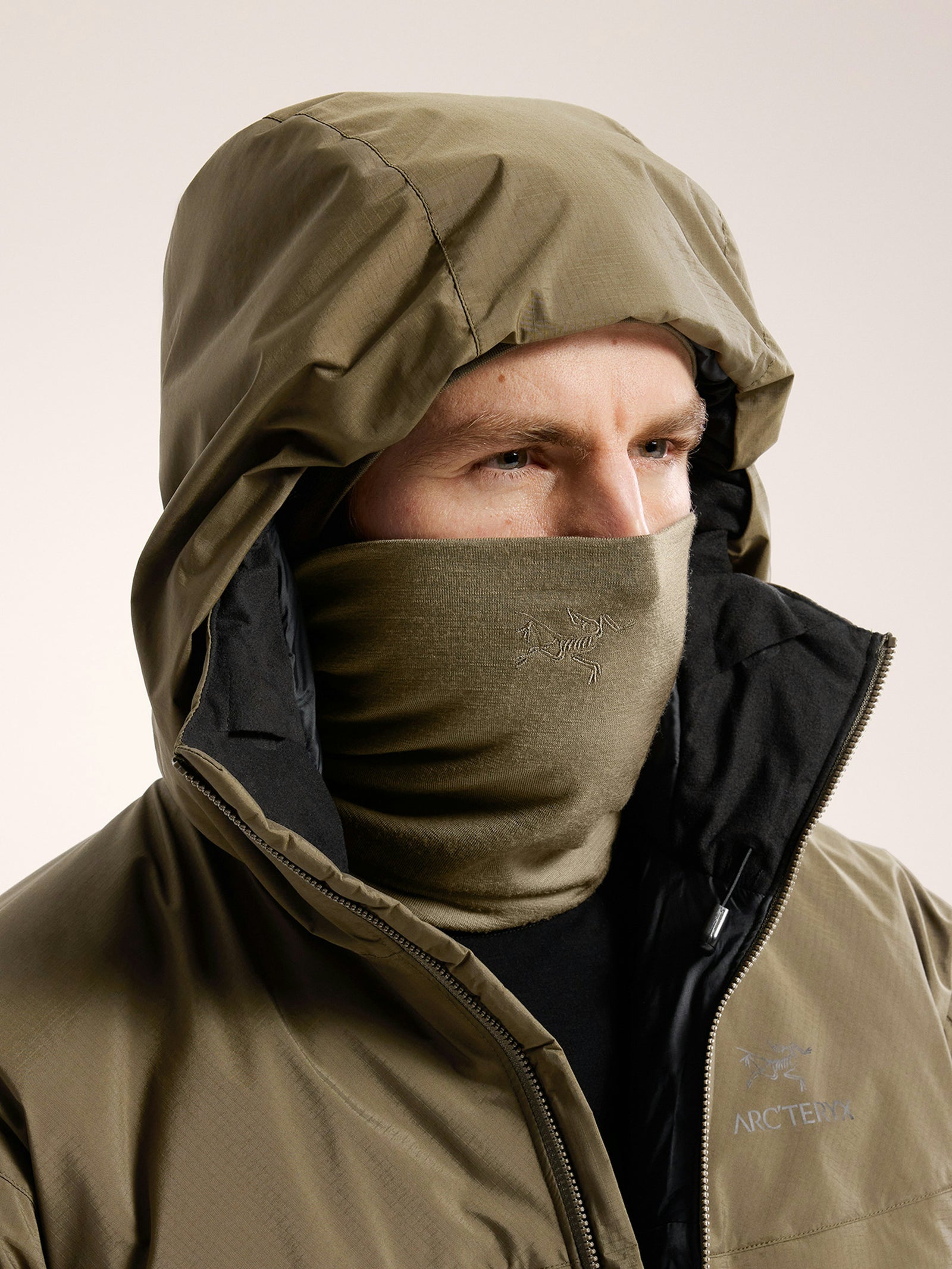 Eine Person trägt eine olivgrüne Kapuzenjacke und die ARC'TERYX LEAF Arc'teryx Cold WX Neck Gaiter AR, die den Hals und das untere Gesicht bedeckt und so einen effektiven Schutz vor kaltem Wetter bietet - nur Augen und Nase sind sichtbar.