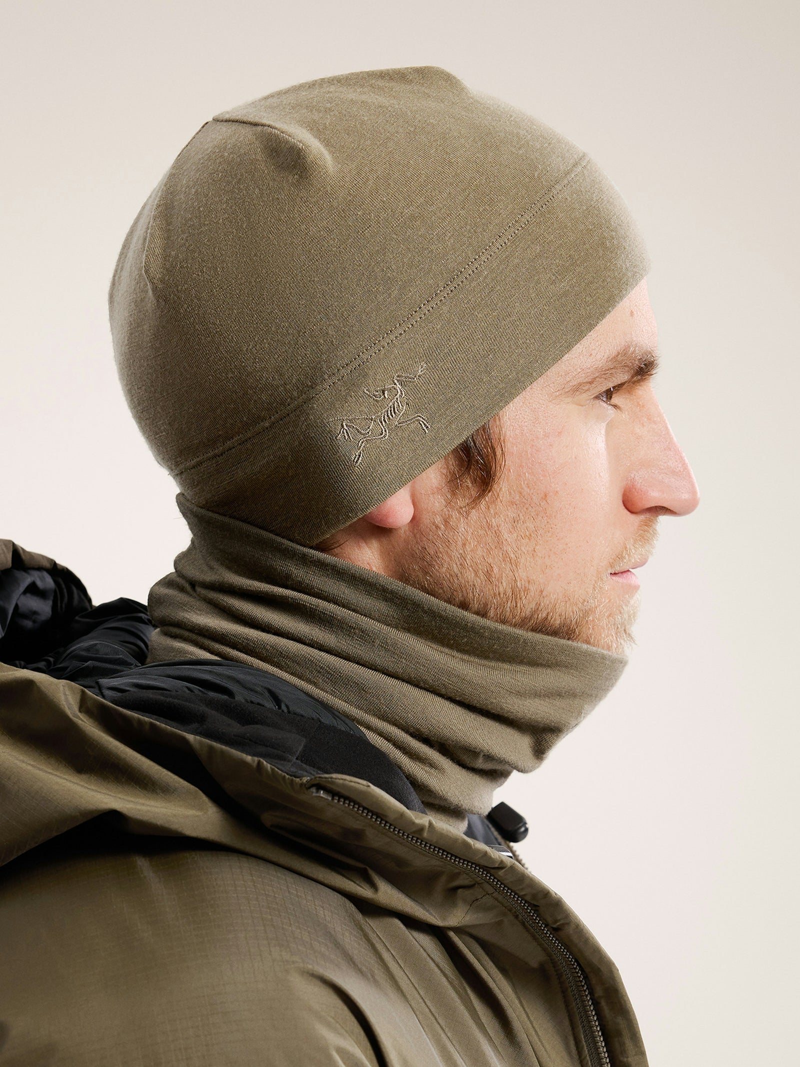 Ein Mann im Profil trägt eine hellolivfarbene ARC'TERYX LEAF Arc'teryx Cold WX Neck Gaiter AR und eine passende Mütze zu einer grünen Jacke. Ein schlichter, neutraler Hintergrund unterstreicht sein wintertaugliches, schützendes Outfit.