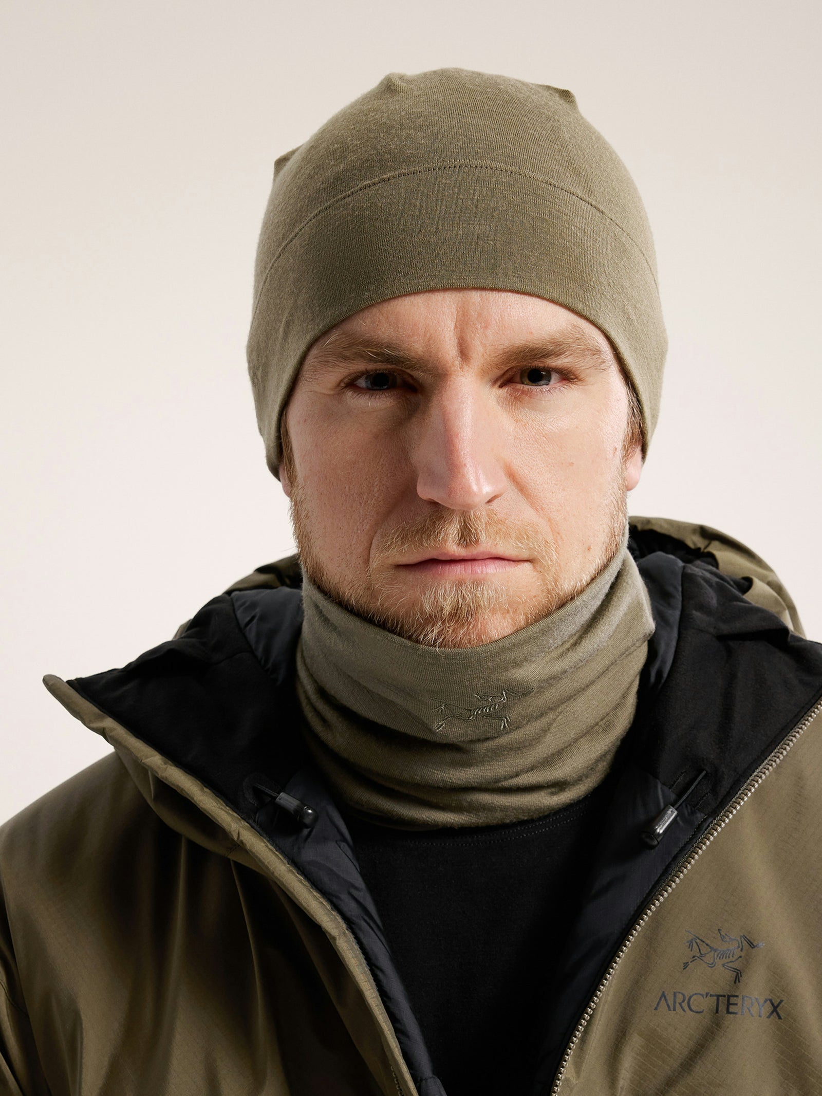 Ein Mann mit olivfarbener Mütze, Arc'teryx Cold WX Neck Gaiter AR by ARC'TERYX LEAF für optimalen Kälteschutz und einer dunkelgrünen Arcteryx-Jacke schaut mit neutralem Gesichtsausdruck direkt in die Kamera.