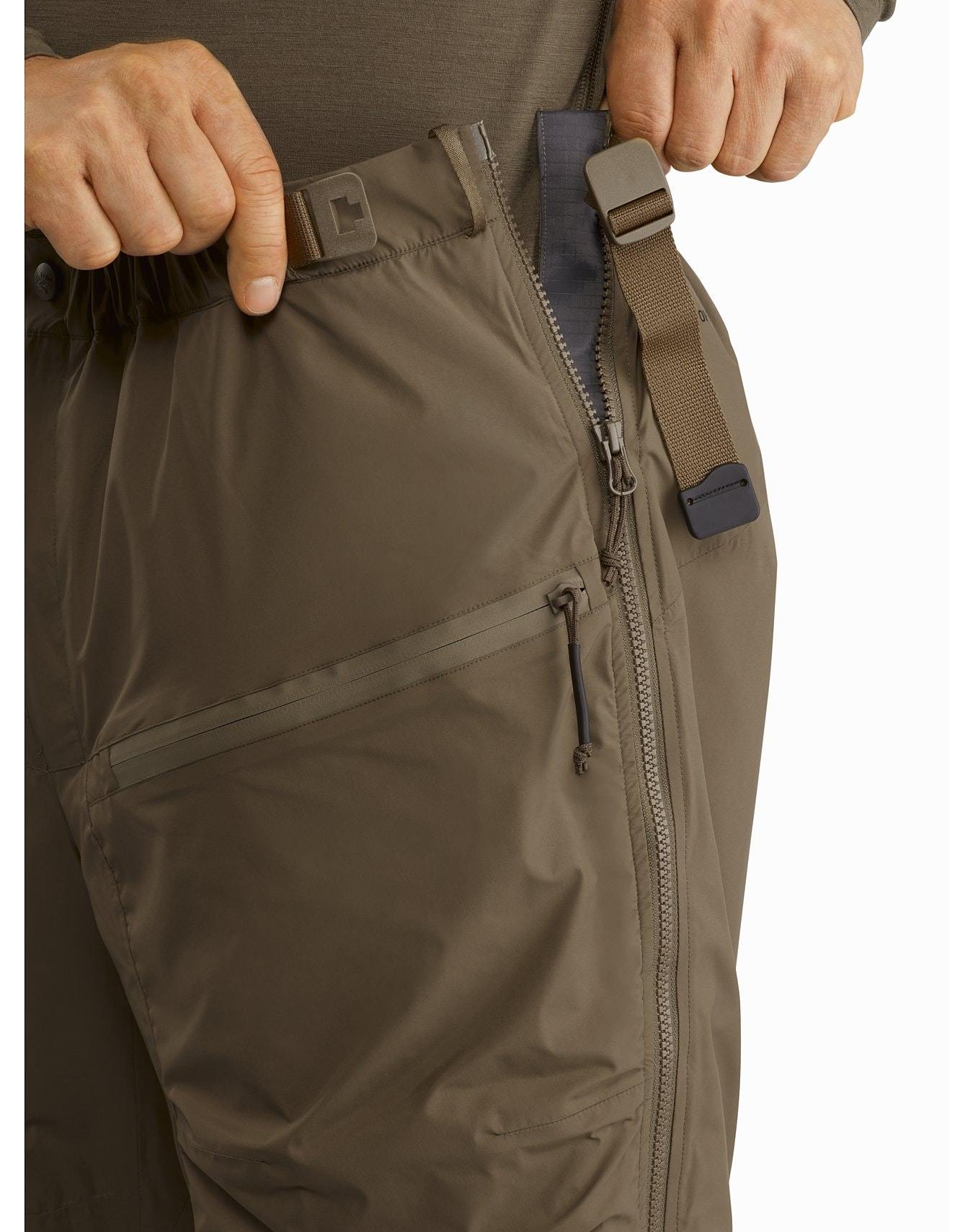 Eine Person verstellt den Bund der schwarzen Arc'teryx Cold WX Pant LT Gen2 von ARC'TERYX LEAF, die mit einer Reißverschlusstasche, einem Reißverschluss und einem verstellbaren Riemen mit Kunststoffschnalle für optimalen Sitz und Komfort ausgestattet ist.