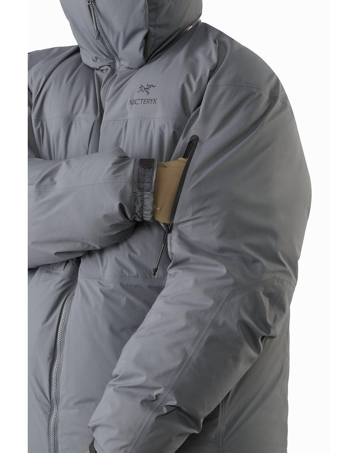 Eine Person in einem grauen ARC'TERYX LEAF Arc'teryx Cold WX Parka SVX - Wolf steht mit verschränkten Armen da. An der oberen Ärmeltasche sind ein brauner Aufnäher und ein Stift befestigt. Die isolierte Herrenjacke ist mit Reißverschluss und Kapuze geschlossen.