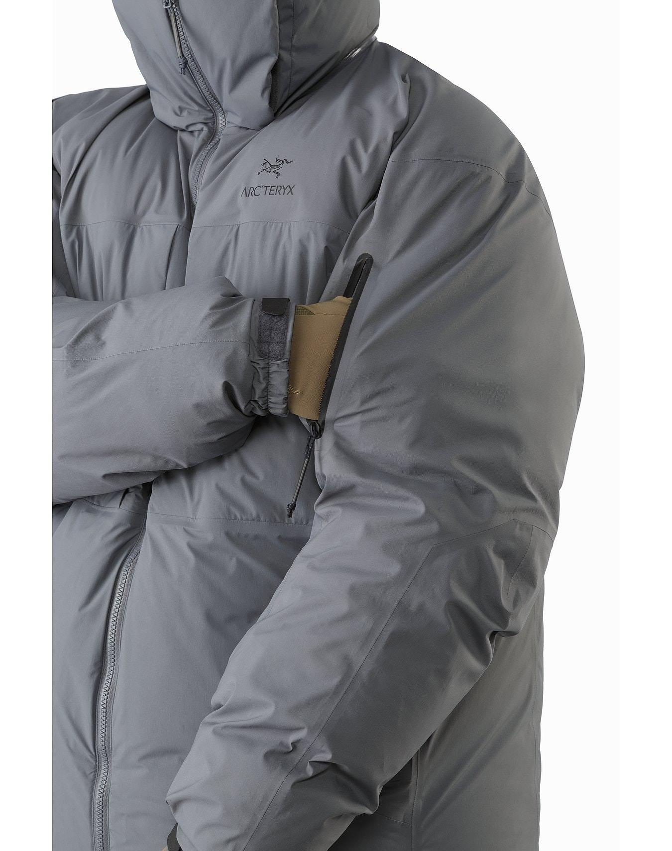 Eine Person in einem grauen ARC'TERYX LEAF Arc'teryx Cold WX Parka SVX - Wolf steht mit verschränkten Armen da. An der oberen Ärmeltasche sind ein brauner Aufnäher und ein Stift befestigt. Die isolierte Herrenjacke ist mit Reißverschluss und Kapuze geschlossen.