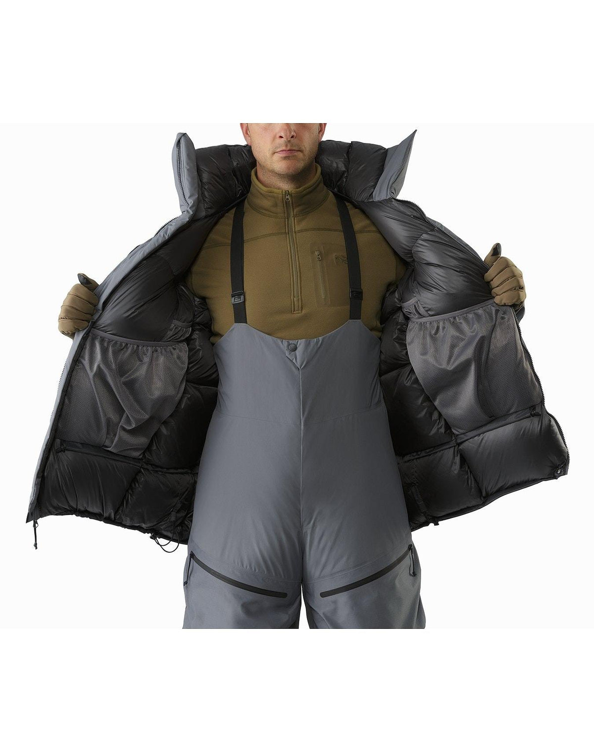 Eine Person in einem braunen Pullover, einem grauen isolierten Latz, Handschuhen und einem ARC'TERYX LEAF Cold WX Parka SVX - Wolf hält die Jacke offen, um große Innentaschen zu zeigen. Das Gesicht ist teilweise aus dem Rahmen.
