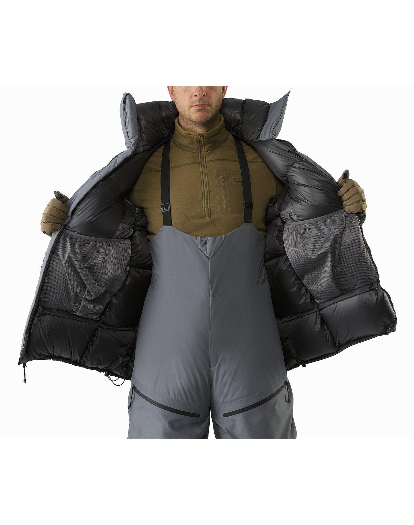 Eine Person in einem braunen Pullover, einem grauen isolierten Latz, Handschuhen und einem ARC'TERYX LEAF Cold WX Parka SVX - Wolf hält die Jacke offen, um große Innentaschen zu zeigen. Das Gesicht ist teilweise aus dem Rahmen.