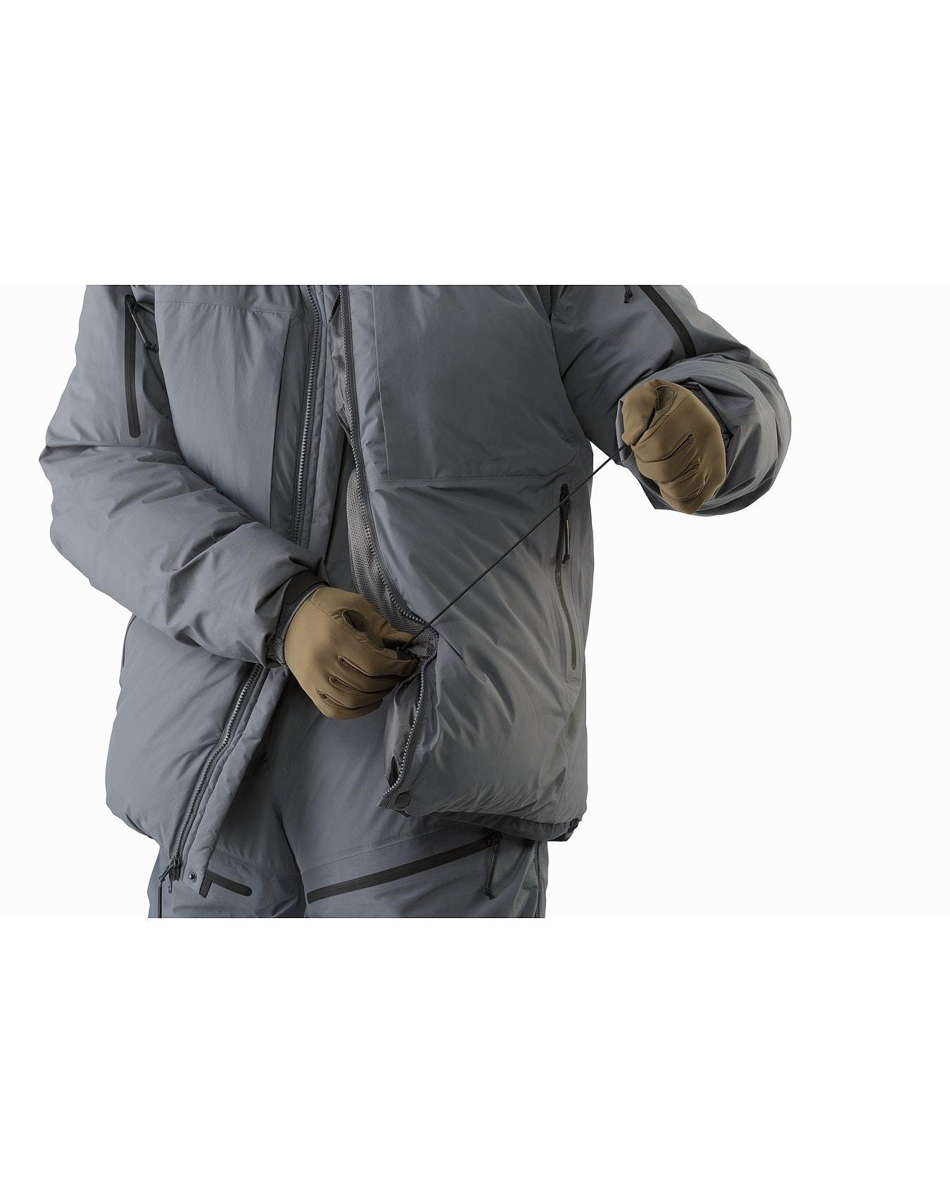 Eine Person mit braunen Handschuhen zieht den Taillenkordelzug eines ARC'TERYX LEAF Arc'teryx Cold WX Parka SVX - Wolf zu, wobei nur ihr Oberkörper und ihre Arme vor einem weißen Hintergrund zu sehen sind.