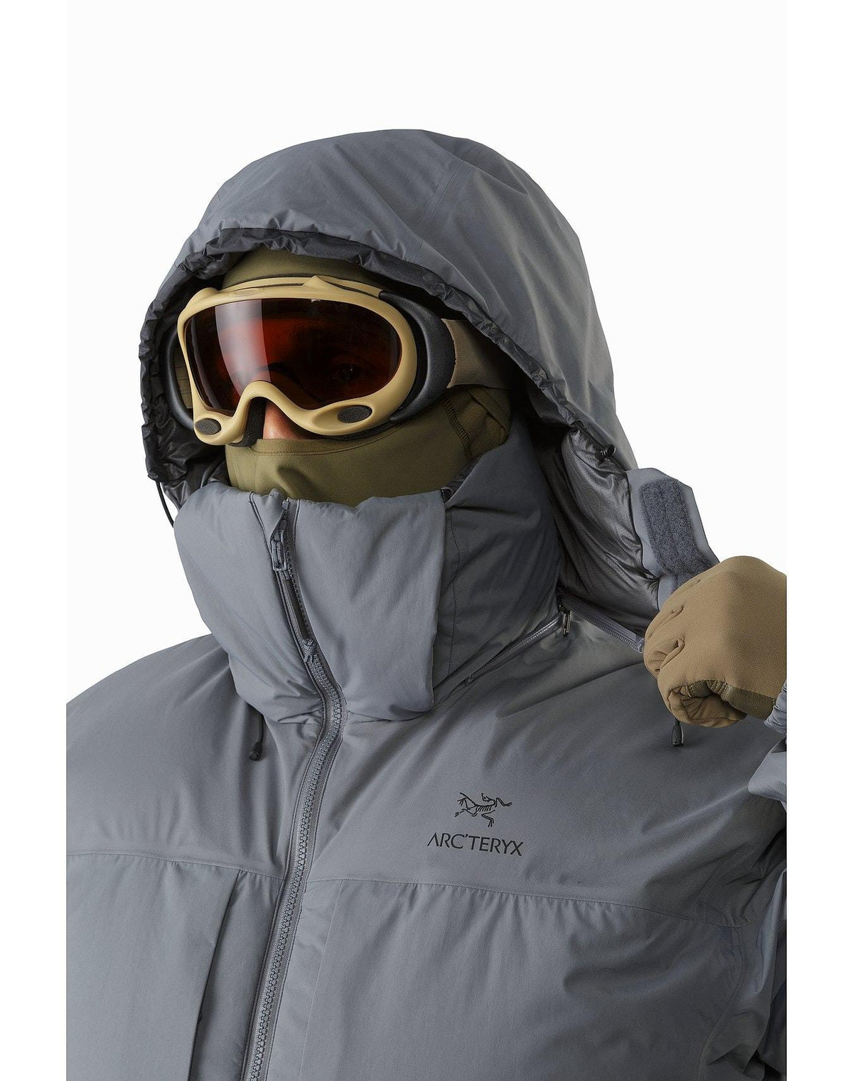 Eine Person in einem grauen ARC'TERYX LEAF Arc'teryx Cold WX Parka SVX - Wolf mit hochgezogener Kapuze, beigen Handschuhen, Skibrille und einer Gesichtsbedeckung verstellt die Kapuze. Diese isolierte Herrenjacke ist ideal für kaltes Wetter oder Wintersport.