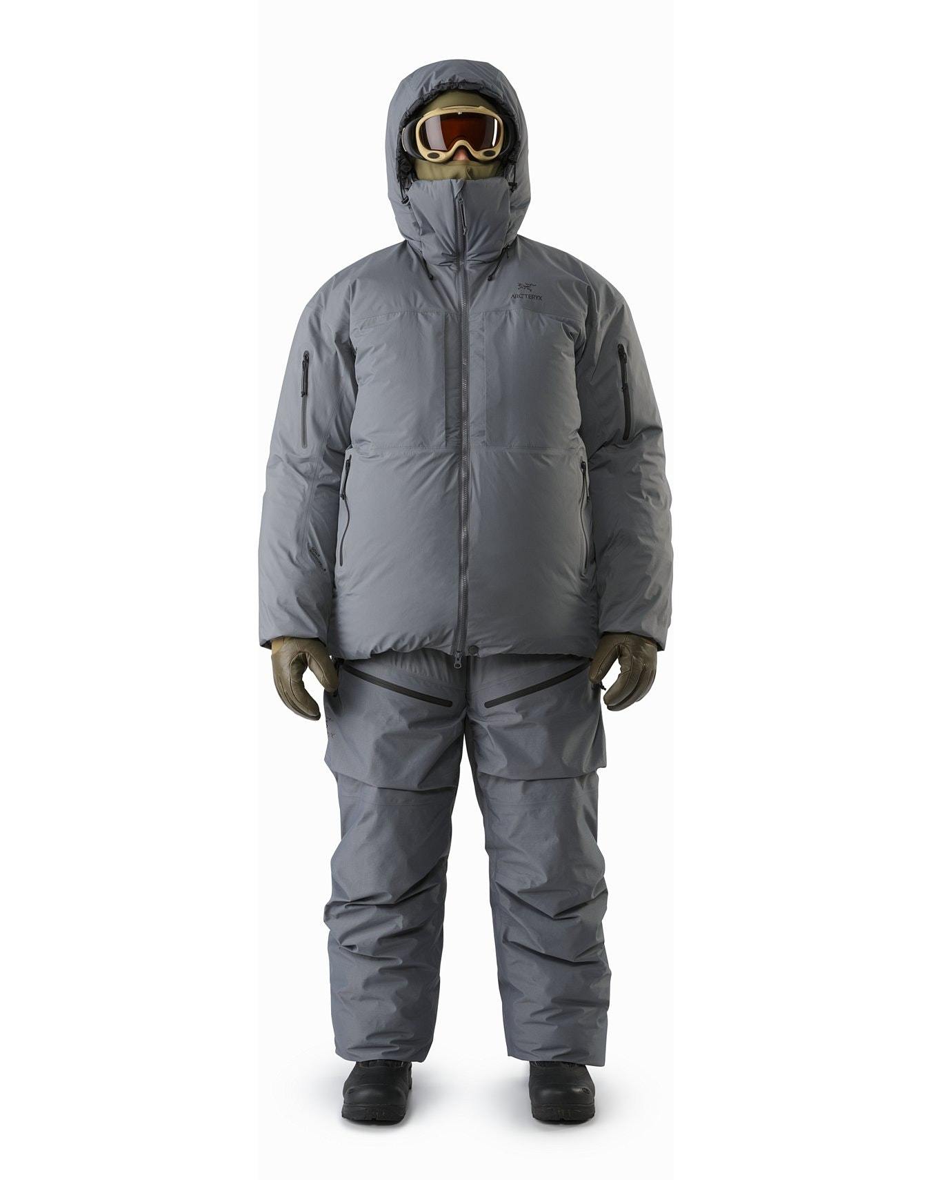 Eine Person steht mit dem Blick nach vorne und trägt den ARC'TERYX LEAF Arc'teryx Cold WX Parka SVX - Wolf, mit passender isolierter Hose, Handschuhen, dunklen Stiefeln und gelber Skibrille - bereit für verschneite oder kalte Wetterbedingungen.