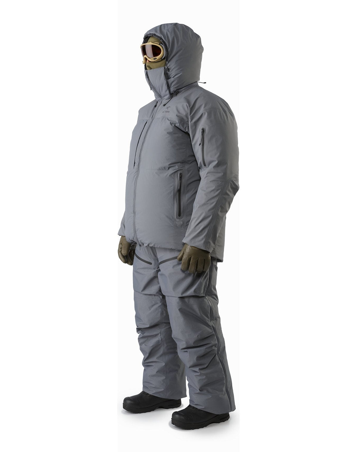 Eine Person in einem grauen ARC'TERYX LEAF Arc'teryx Cold WX Parka SVX - Wolf, isolierter Winterhose, braunen Handschuhen, schwarzen Stiefeln, Schutzbrille und Kapuze steht leicht zur Seite gewandt.