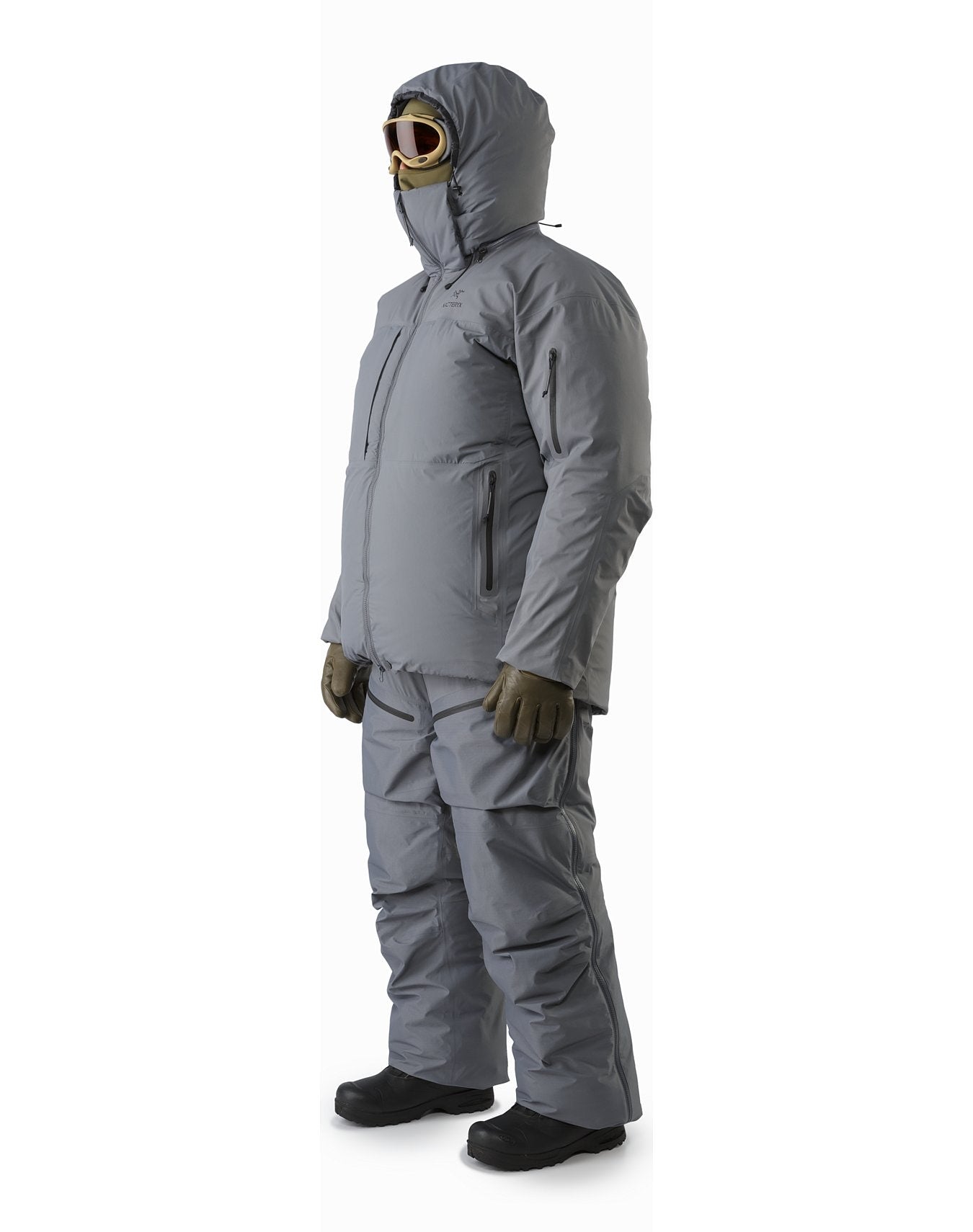 Eine Person in einem grauen ARC'TERYX LEAF Arc'teryx Cold WX Parka SVX - Wolf, isolierter Winterhose, braunen Handschuhen, schwarzen Stiefeln, Schutzbrille und Kapuze steht leicht zur Seite gewandt.
