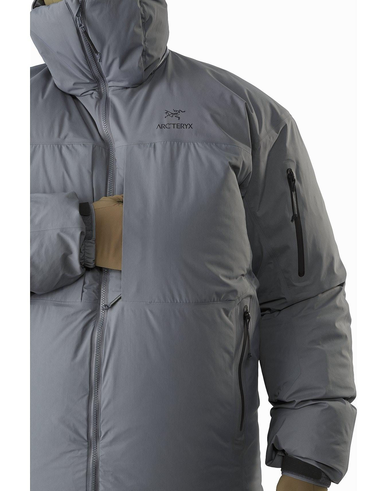 Wenn man den ARC'TERYX LEAF Arc'teryx Cold WX Parka SVX - Wolf mit hochgezogener Kapuze und hellbraunen Handschuhen trägt, steckt man die Hand in die Brusttasche. Die daunenisolierte Herrenjacke hat mehrere Reißverschlusstaschen und einen hohen Kragen.