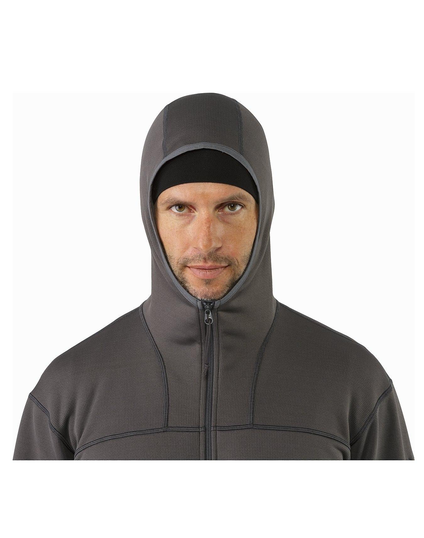 Eine Person trägt einen ARC'TERYX LEAF Arc'teryx Naga Hoody Full Zip Gen2.1 in Crocodile mit hochgezogener Kapuze, die den Kopf und den größten Teil der Stirn bedeckt, und schaut direkt in die Kamera vor einem weißen Hintergrund.