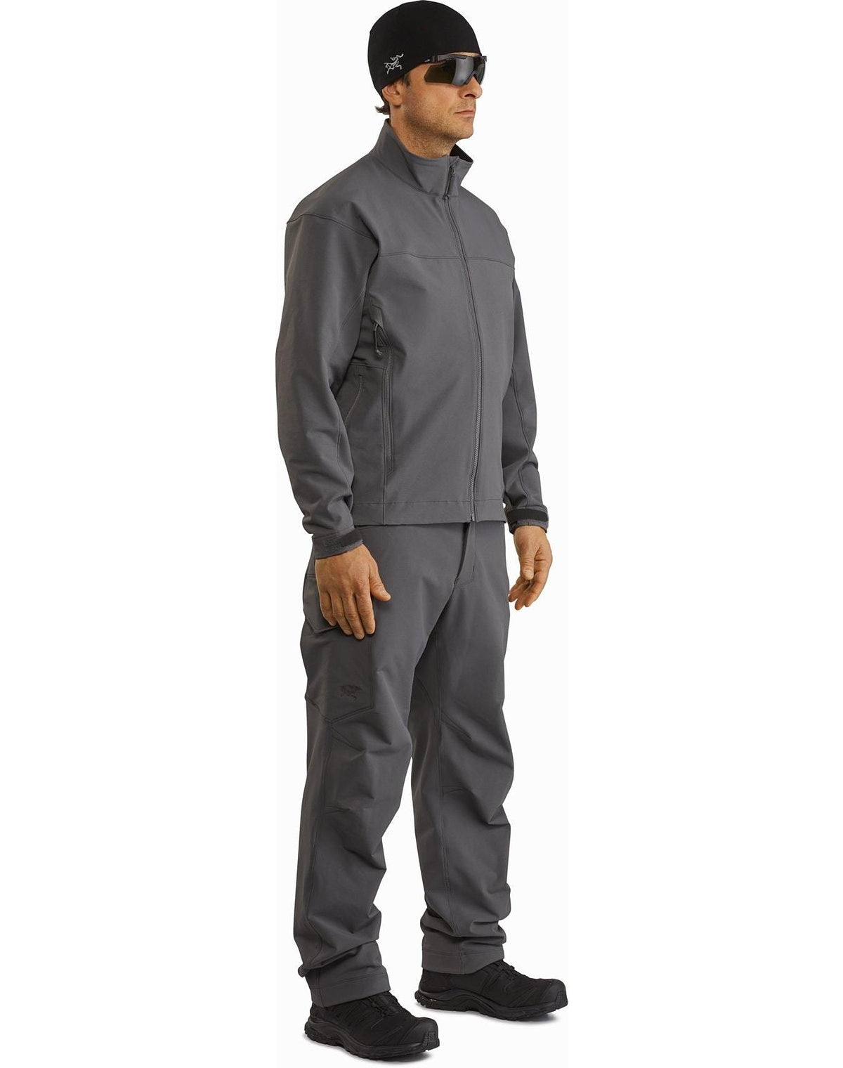 Ein Mann steht in einer Ranger Green Arc'teryx Patrol Jacket AR Men's von ARC'TERYX LEAF mit passender Hose, schwarzen Schuhen, Sonnenbrille und einer schwarzen Mütze. Er blickt leicht zur Seite und hält die Hände entspannt an den Seiten vor einem weißen Hintergrund.