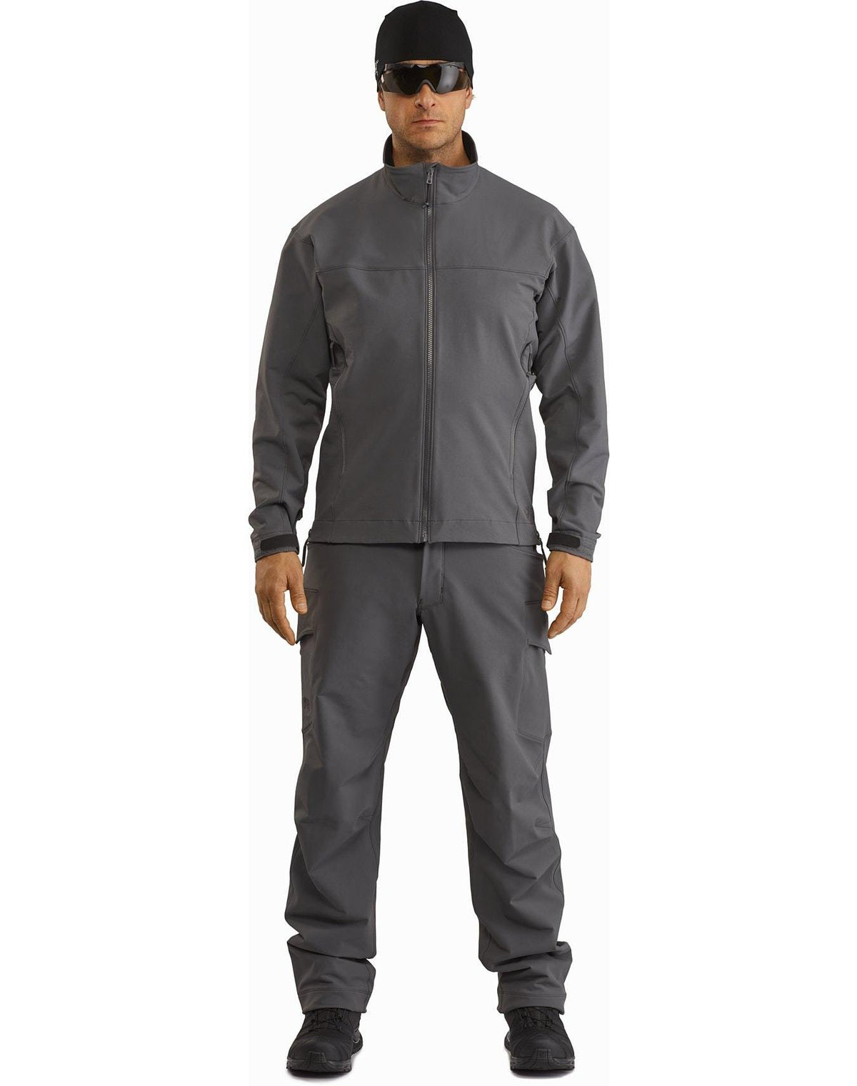 Eine Person steht mit dem Gesicht nach vorne und trägt eine ARC'TERYX LEAF Arc'teryx Patrol Jacket AR Men's - Wolf mit passender Hose, schwarzen Schuhen, einer dunklen Sonnenbrille und einer schwarzen Kopfbedeckung vor einem schlichten weißen Hintergrund.