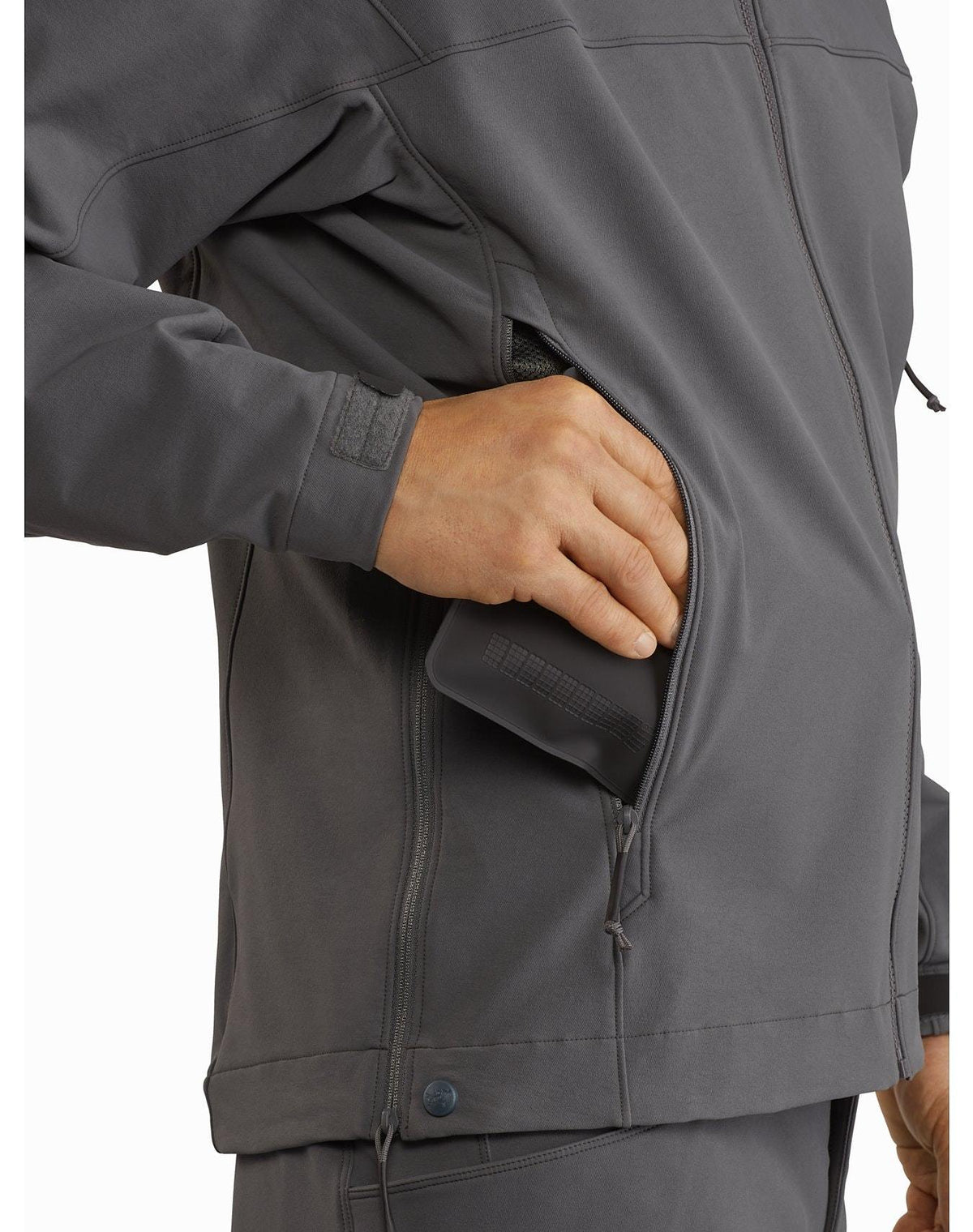 Eine Person, die die ARC'TERYX LEAF Arc'teryx Patrol Jacket AR Men's - Wolf trägt, steckt ein Smartphone in die Seitentasche mit Reißverschluss. Nur die Hand und ein Teil des Oberkörpers in der Jacke sind sichtbar.