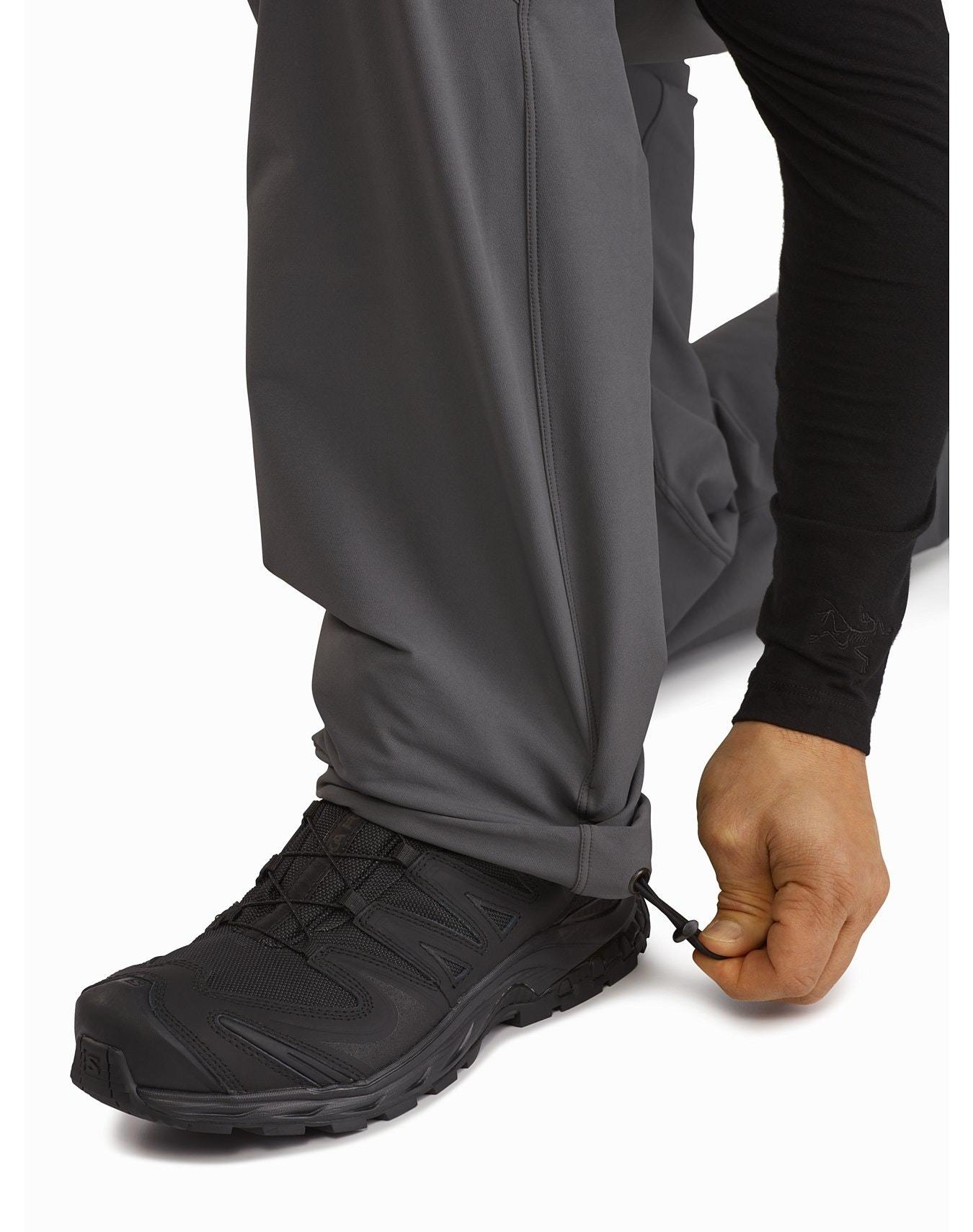 Eine Person kniet, um den Kordelzug am Knöchel zu regulieren und das Bündchen der schwarzen Arc'teryx Patrol Pant AR Men's von ARC'TERYX LEAF zu schließen, während sie schwarze Trail-Schuhe trägt.