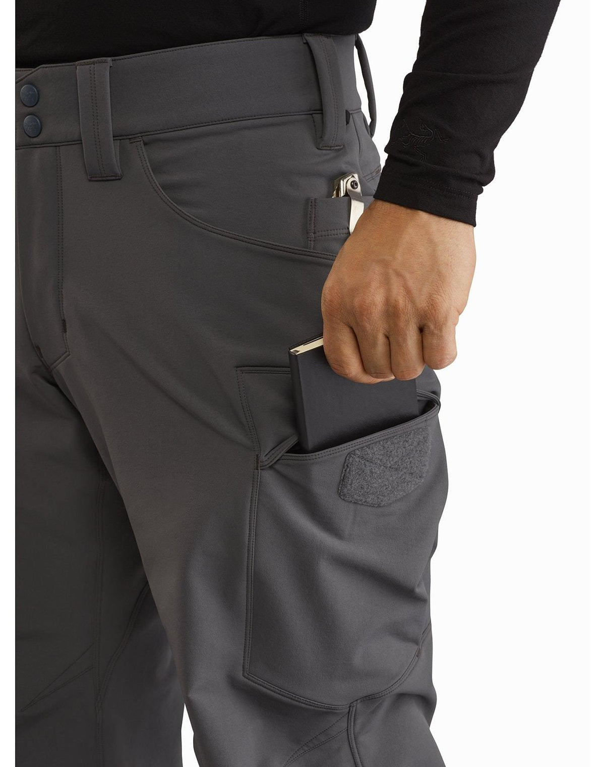 Eine Person, die die ARC'TERYX LEAF Arc'teryx Patrol Pant AR Men's - Black trägt, steckt ein schwarzes Portemonnaie in eine Oberschenkeltasche mit Klettverschluss und konzentriert sich dabei auf die Hand und die Tasche.