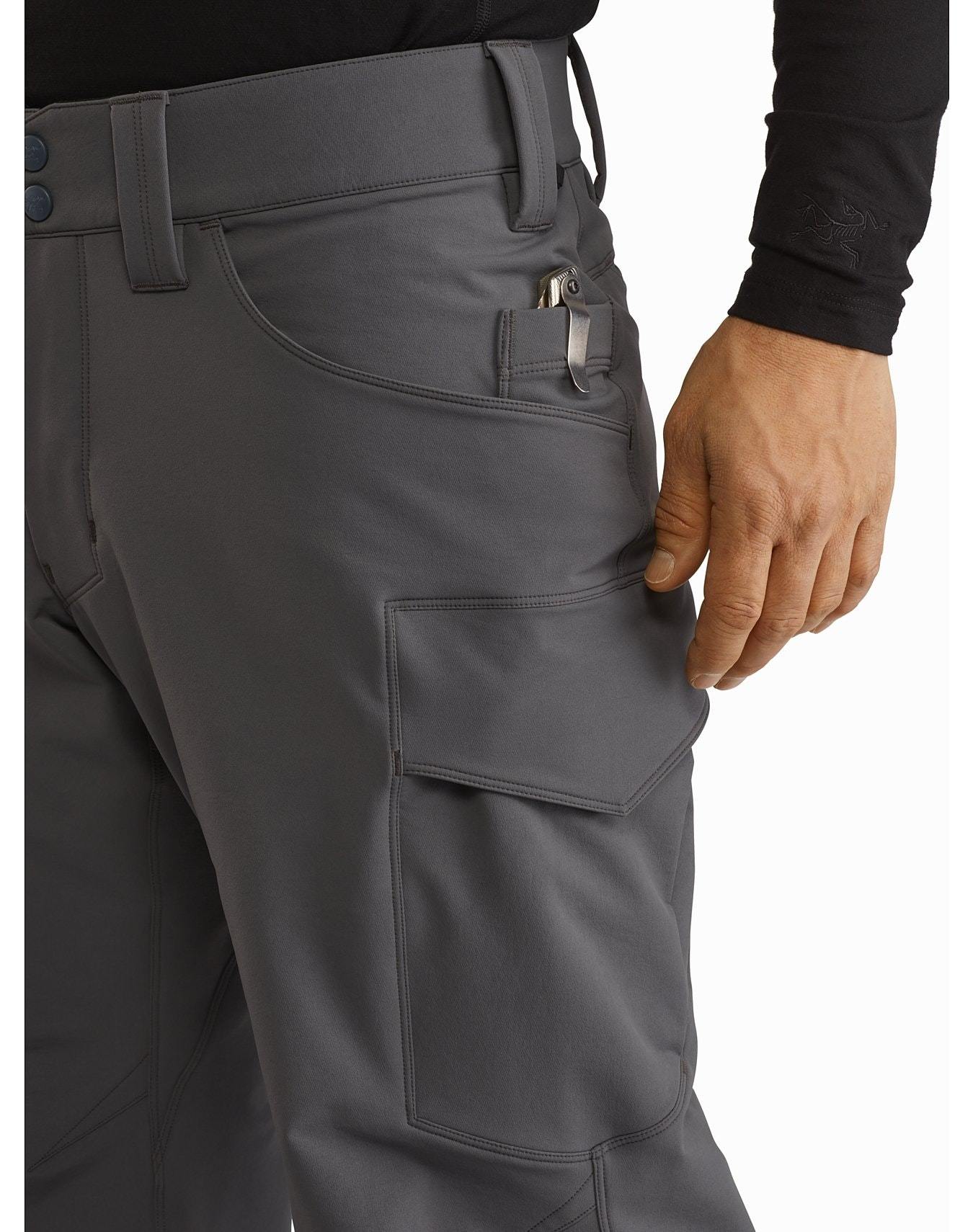 Eine Person, die eine ARC'TERYX LEAF Arc'teryx Patrol Pant AR Men's - Wolf Cargohose mit mehreren Taschen und einem kleinen Werkzeug, das an einer Vordertasche befestigt ist, trägt, steht mit der rechten Hand in der Nähe einer Oberschenkeltasche.