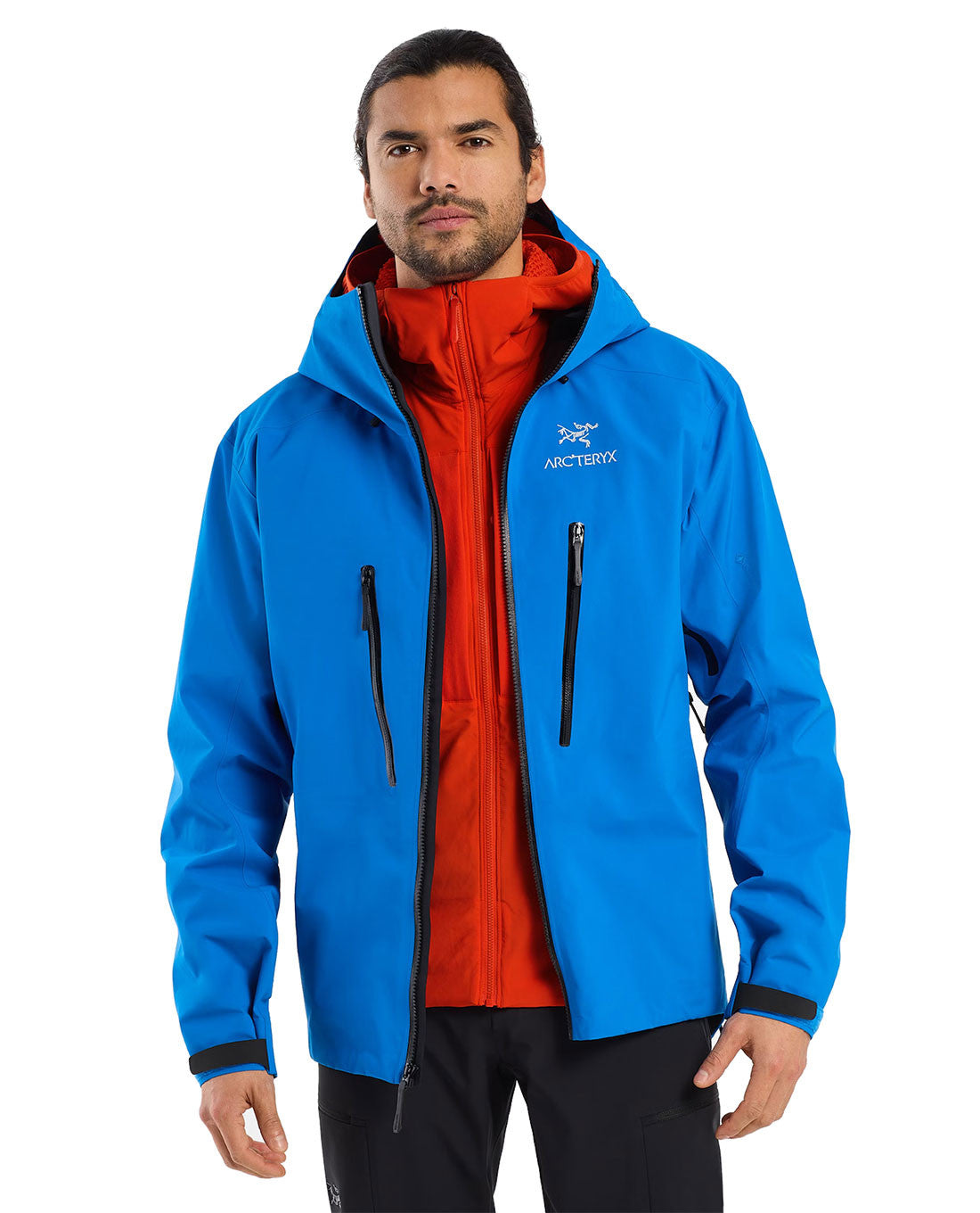 Ein Mann mit langen dunklen Haaren und einem Bart trägt eine leuchtend blaue ARC'TERYX PRO Arc'teryx Practitioner AR Hoody Men's - Dynasty, eine atmungsaktive Zwischenjacke mit synthetischer Isolierung, über einer roten Jacke mit Reißverschluss. Er blickt nach vorne und hat einen neutralen Ausdruck.