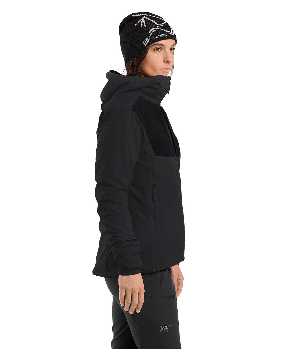 Eine Person steht im Profil und trägt den ARC'TERYX PRO Arc'teryx Practitioner AR Hoody Women's - Carbon Copy, eine passende schwarze Hose und eine schwarze Mütze mit weißen Akzenten. Der atmungsaktive Kapuzenpullover mit Kapuze ist vor einem schlichten weißen Hintergrund zu sehen.