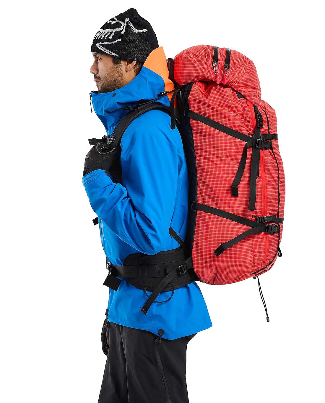Eine Person in blauer Jacke, schwarzen Handschuhen und schwarzer Mütze steht im Profil und trägt den ARC'TERYX PRO Arc'teryx Rescue Pack 50 mit mehreren Riemen und Schnallen vor einem schlichten weißen Hintergrund.