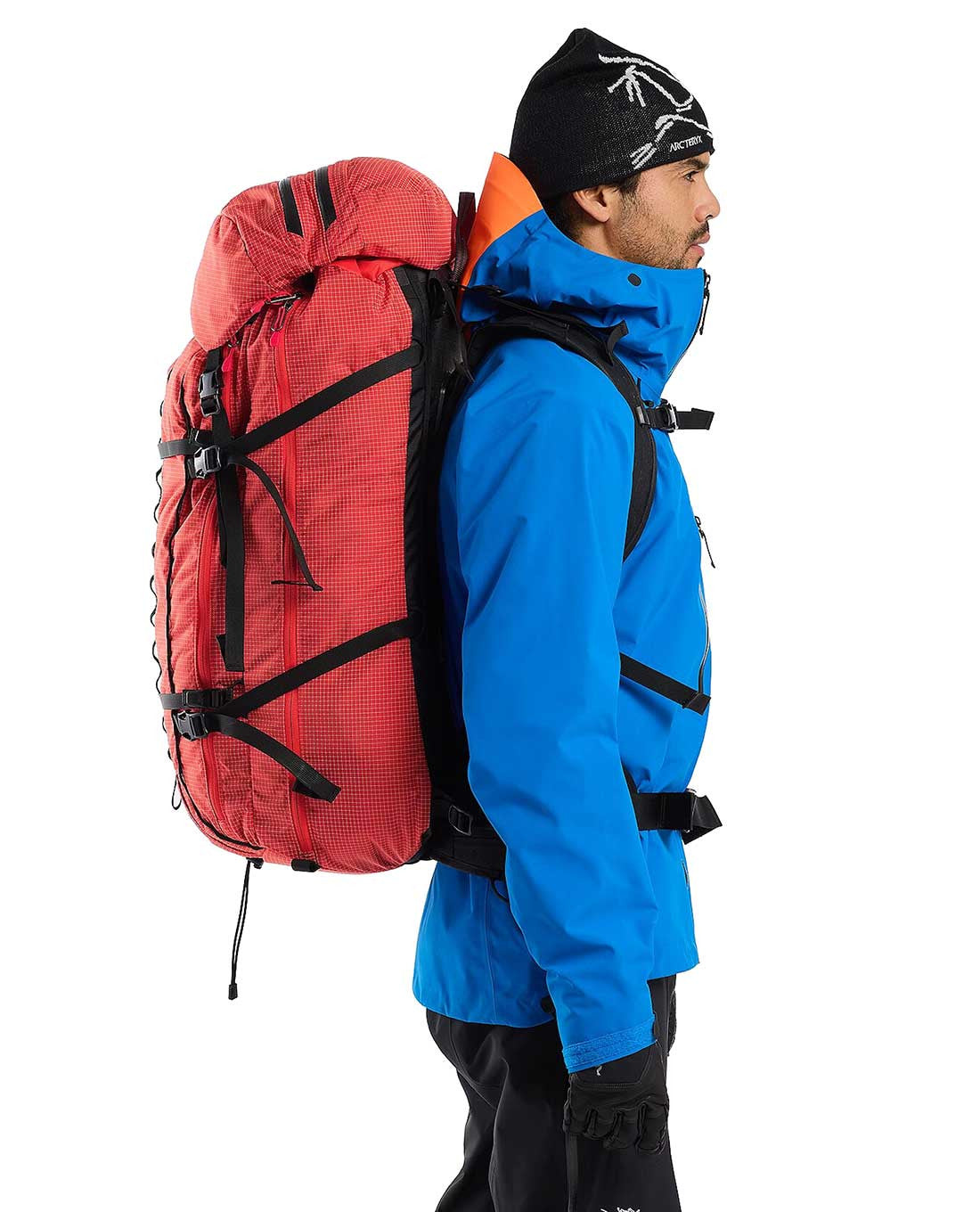 Eine Person mit blauer Jacke, schwarzen Handschuhen und schwarzer Mütze steht im Profil und trägt einen ARC'TERYX PRO Arc'teryx Rescue Pack 50, der mit schwarzen Gurten gesichert ist.