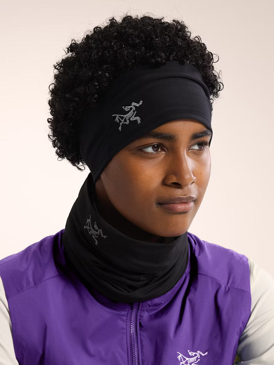 Eine Person mit lockigem Haar trägt ein schwarzes ARC'TERYX Rho Stirnband mit weißem Logo, eine passende Halskrause und eine lila-weiße Jacke vor einem schlichten hellen Hintergrund.