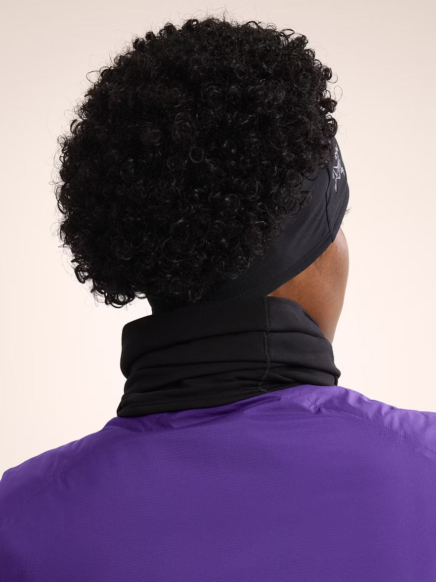 Eine Person mit kurzen, gelockten schwarzen Haaren wird von hinten vor einem hellen Hintergrund gezeigt. Sie trägt ein ARC'TERYX Rho Headband - Black und eine hochgeschlossene schwarz-lila Jacke.
