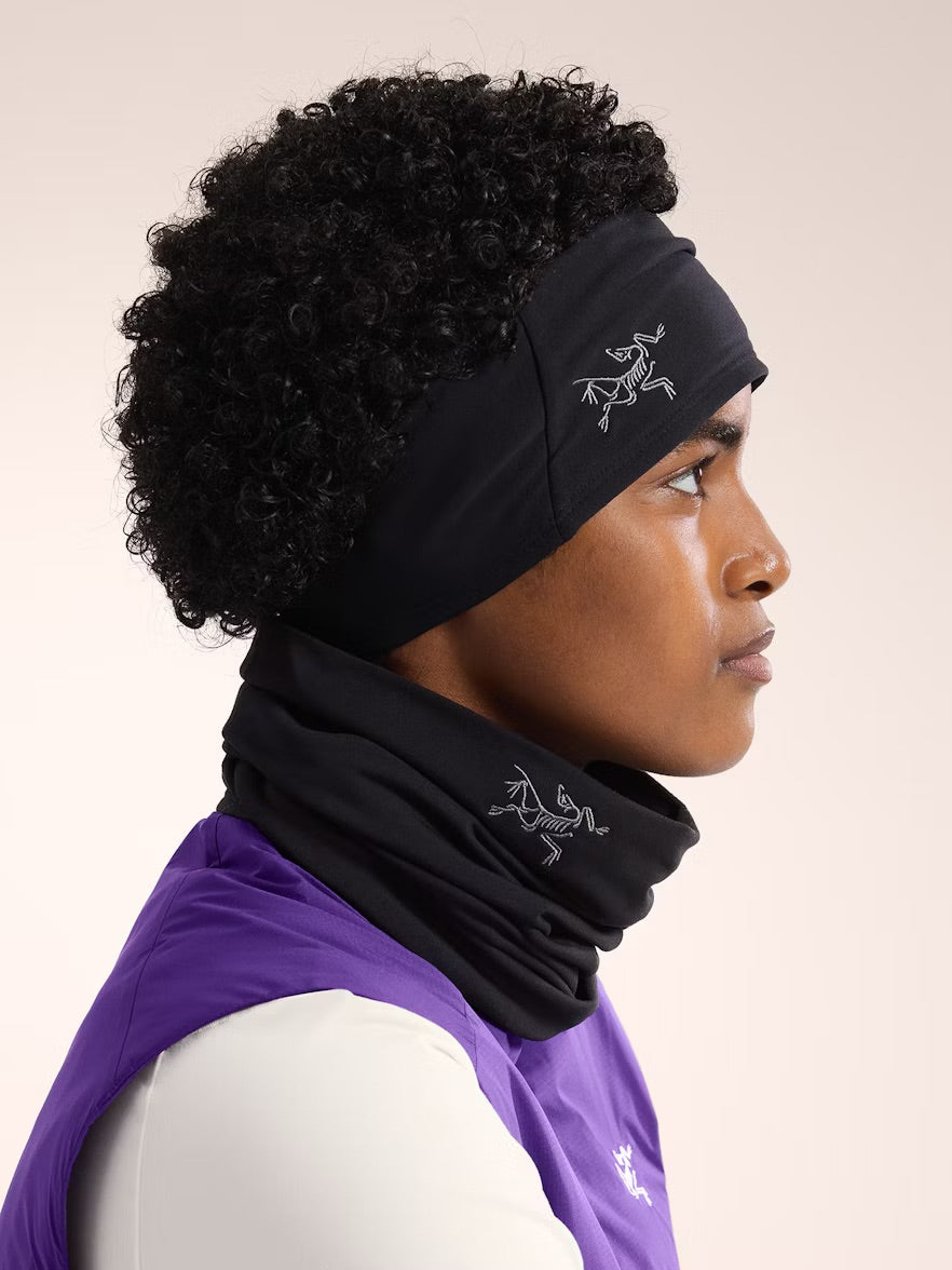 Eine Person mit kurzen lockigen Haaren trägt ein schwarzes Arc'teryx Rho Headband mit weißem Vogellogo, gepaart mit einer Halskrause und einem lila-weißen Oberteil vor einem neutralen Hintergrund.