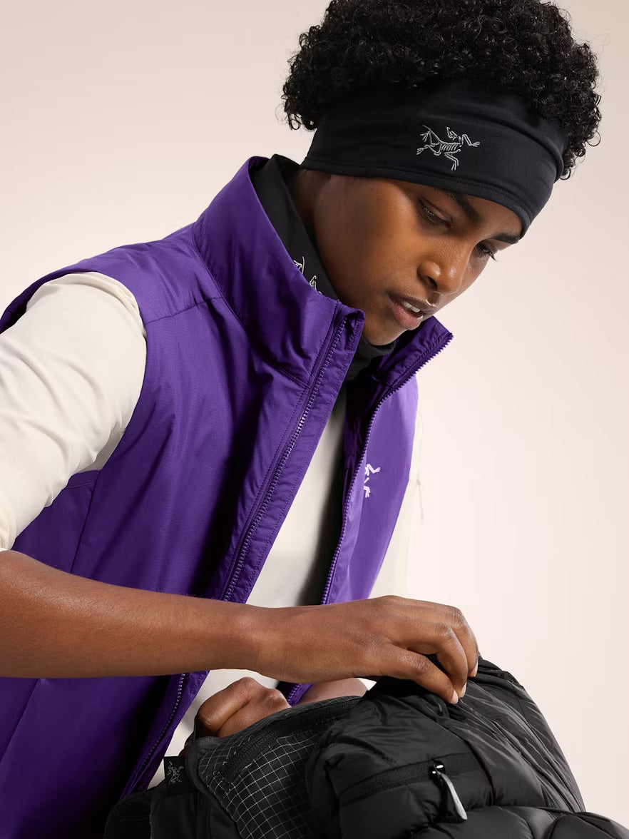 Eine Person mit lockigem Haar trägt das ARC'TERYX Rho Headband - Black und eine lilafarbene Weste. Sie schaut nach unten, während sie eine schwarze Pufferjacke vor einem hellen, einfarbigen Hintergrund zurechtrückt oder einpackt.