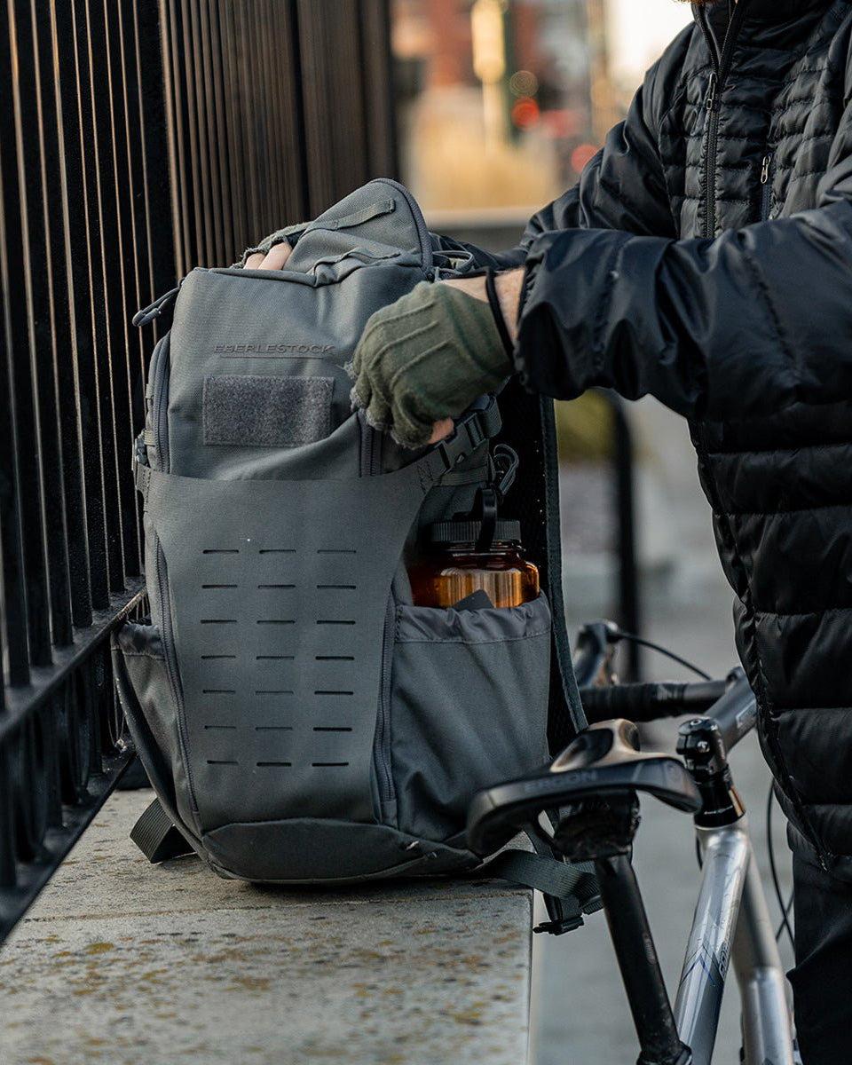 Eine Person, die eine schwarze Jacke und grüne Handschuhe trägt, öffnet einen grauen EBERLESTOCK Bandit Pack mit sichtbarem MOLLE-Panel an einem Fahrrad und einem Metallzaun. In der Seitentasche des Bandit ist eine orangefarbene Flasche sichtbar.