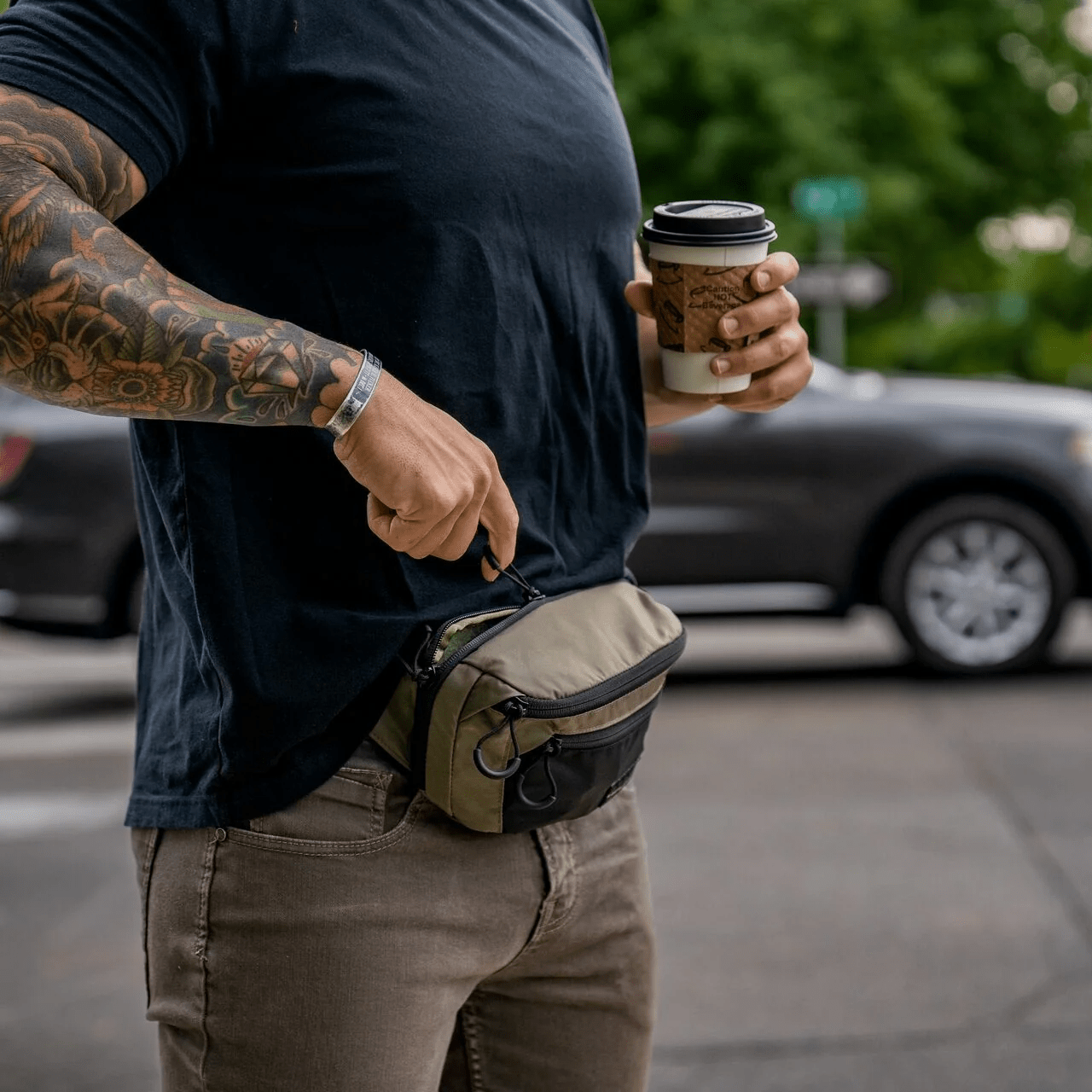 Eine tätowierte Person mit einem Kaffee zum Mitnehmen steht an einem geparkten Auto und öffnet den Reißverschluss einer olivgrünen und schwarzen EBERLESTOCK Bando Bag Standard, die um die Taille getragen wird.