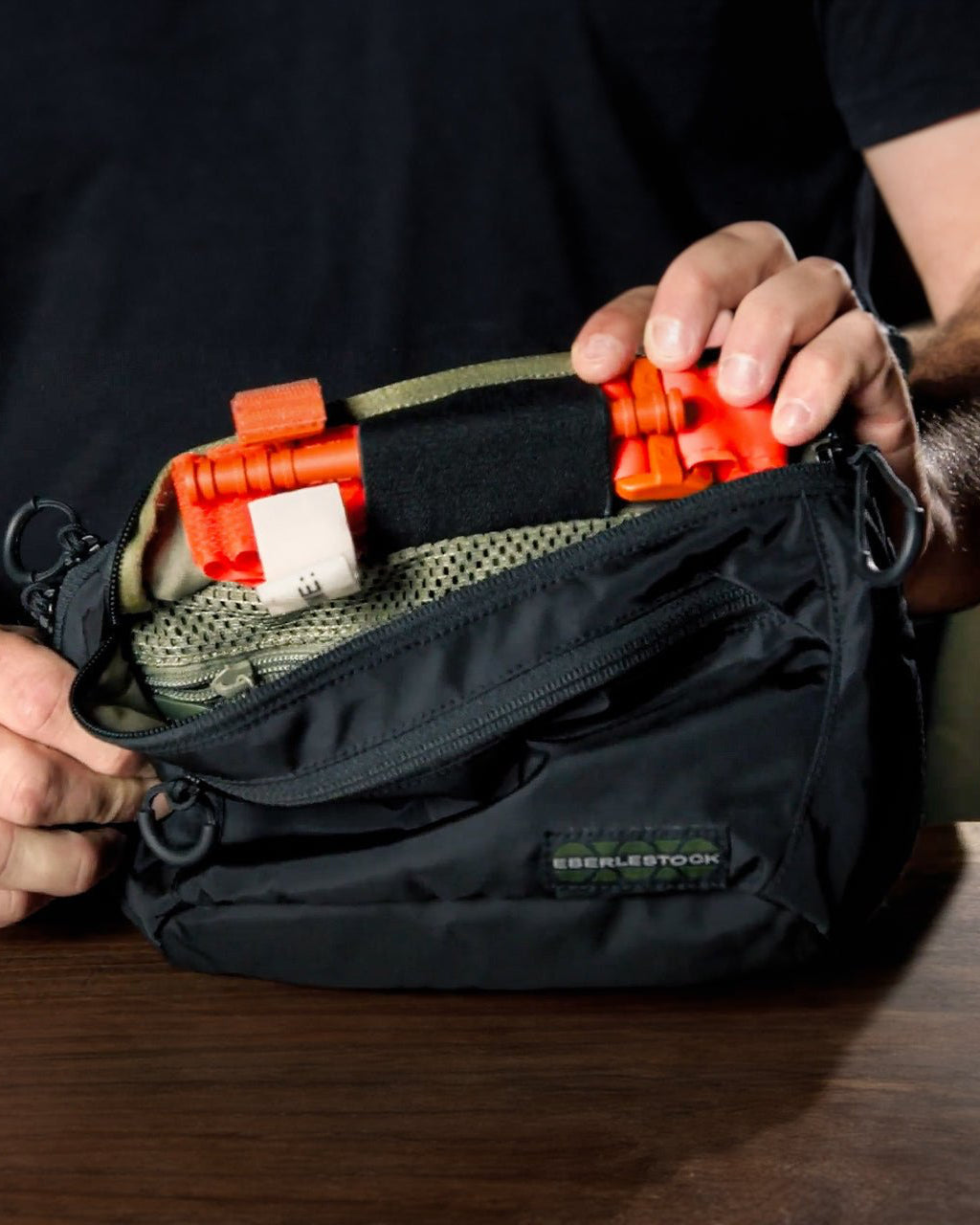 Eine Person öffnet eine kleine schwarze EBERLESTOCK Bando Bag XL auf einem Tisch. Zum Vorschein kommen orangefarbene medizinische Tourniquets und Netztaschen im Inneren - ideal für das verdeckte Tragen in dieser taktischen Umhängetasche.