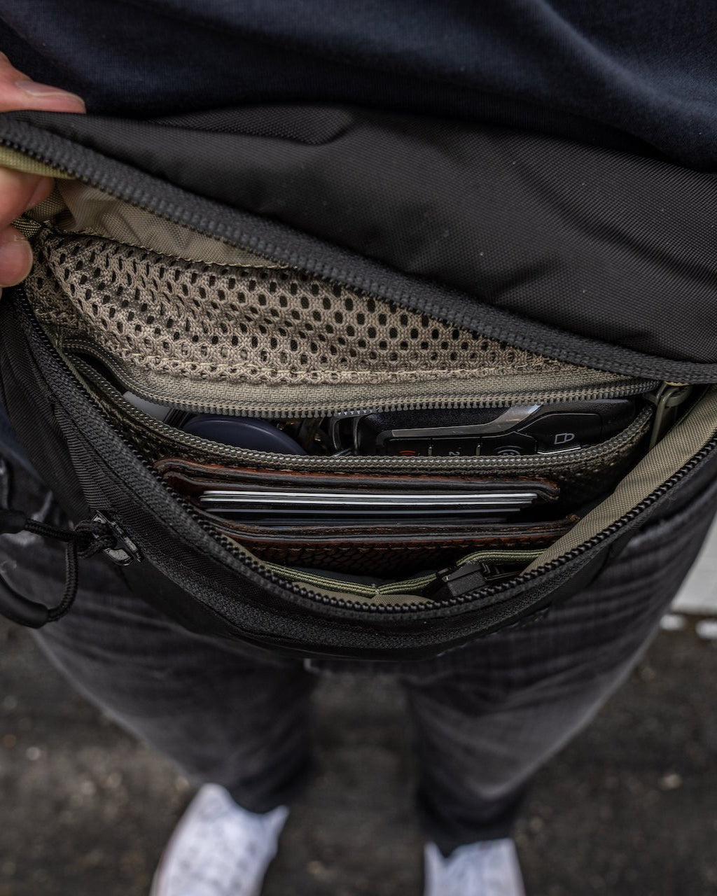 Jemand hält eine EBERLESTOCK Bando Bag XL auf, die sich ideal zum verdeckten Tragen eignet, und zeigt darin ein Telefon, eine Brieftasche und kleine Gegenstände. Der Unterkörper und die weißen Schuhe der Person sind im Hintergrund zu sehen.