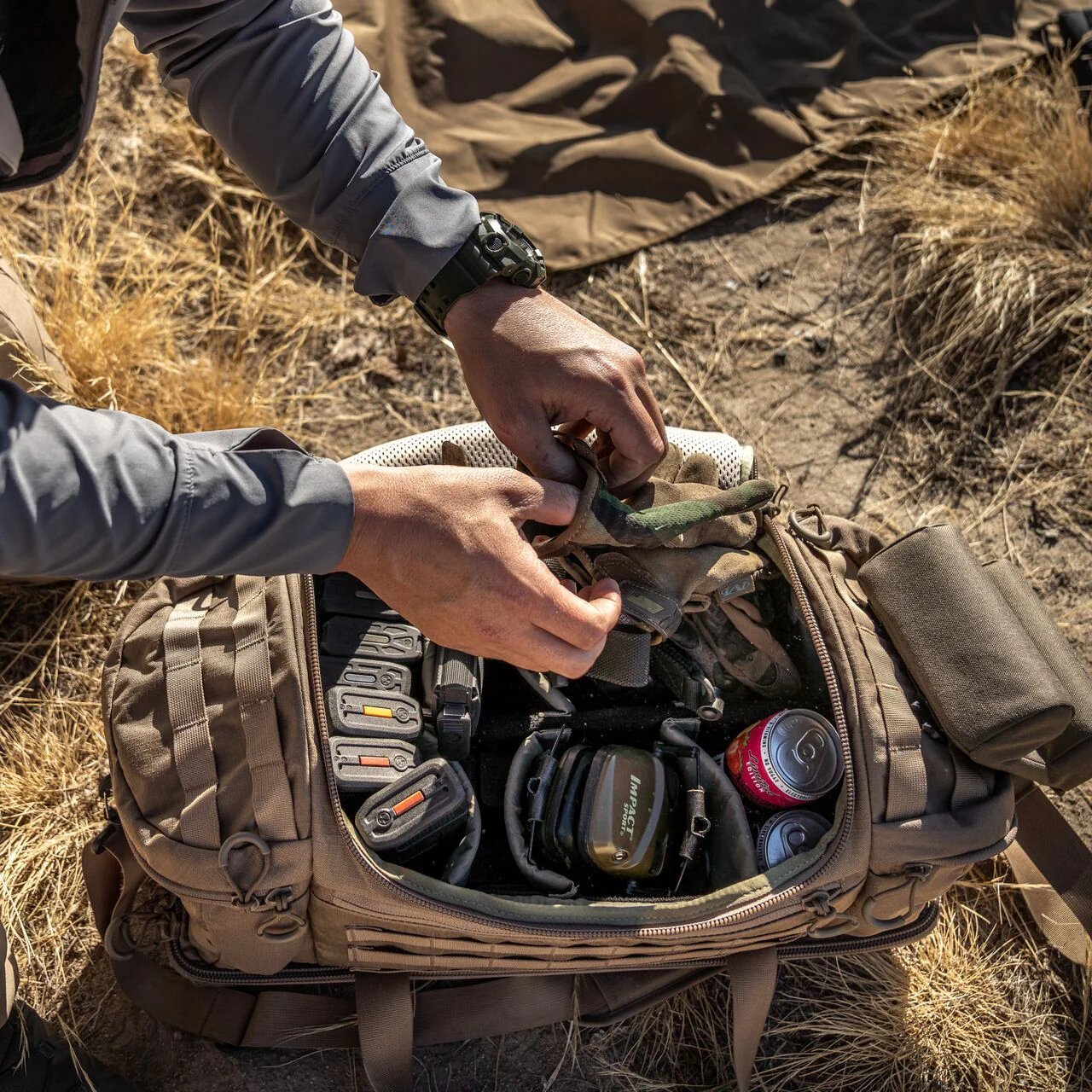 Draußen auf trockenem Gras packt jemand taktische Ausrüstung, Handschuhe, Magazine und eine Limonade in die EBERLESTOCK Eberlestock Bang Bag Range Bag.