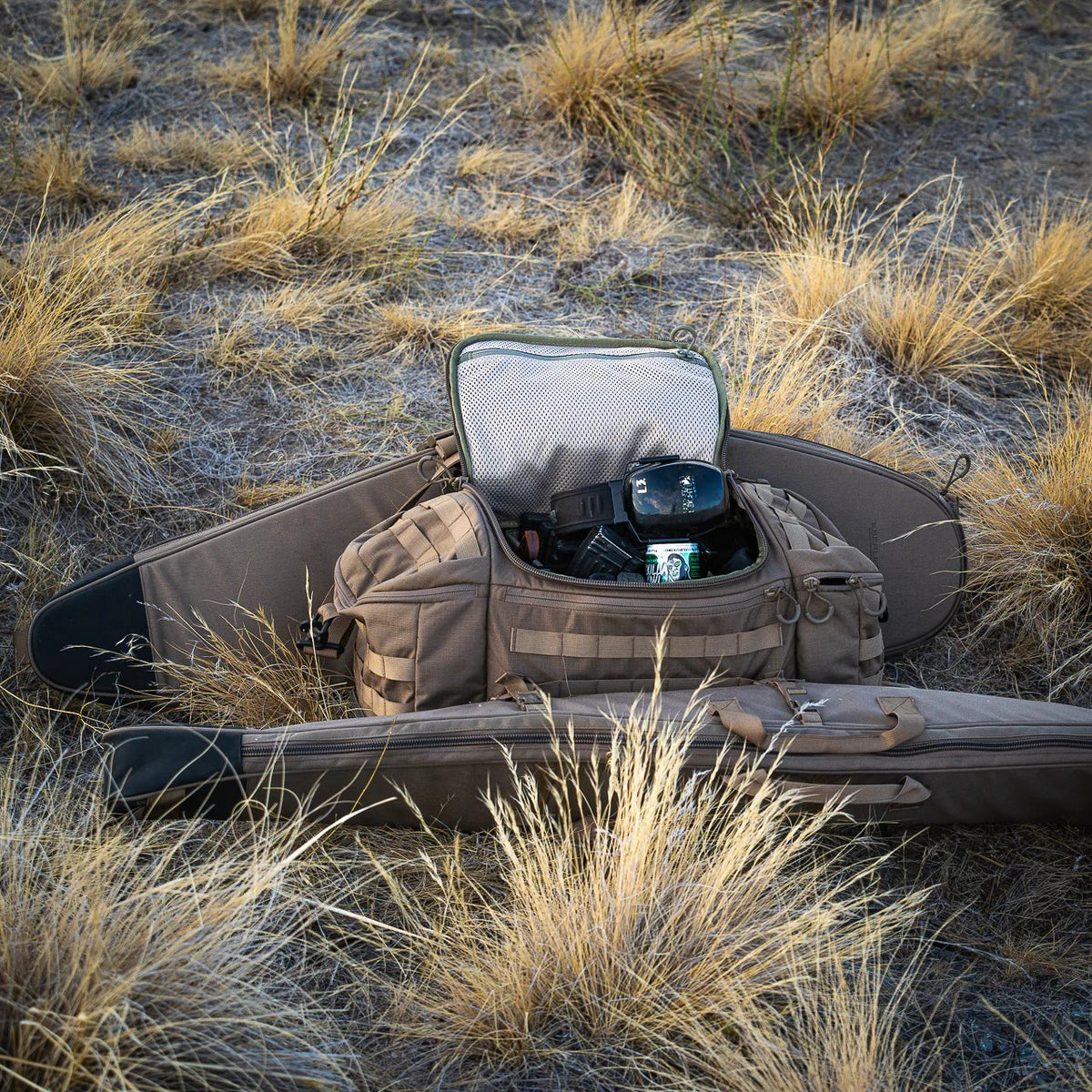 Eine geöffnete EBERLESTOCK Bang Bag zeigt eine Kameraausrüstung und eine Getränkedose, die auf trockenem Gras neben einer geschlossenen Gewehrtasche in einem gelben Feld liegt.