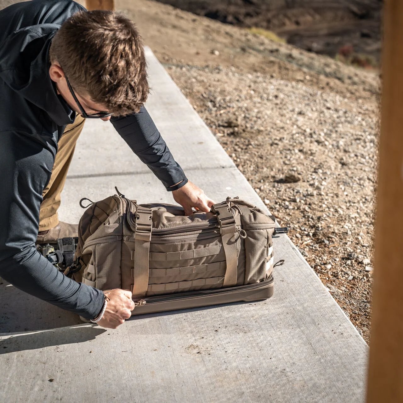 Eine Person mit Sonnenbrille und dunkler Kleidung kniet auf einem Betonweg und öffnet einen EBERLESTOCK Eberlestock Bang Bag, der neben einer sandigen, felsigen Fläche auf dem Boden liegt.