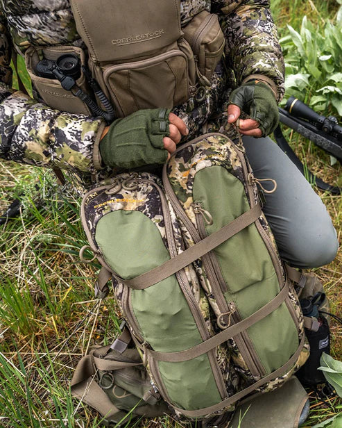 Eine Person in Tarnkleidung kniet im Gras, trägt fingerlose Handschuhe und justiert die Riemen eines EBERLESTOCK Batwing Large - Dry Earth Rucksacks, umgeben von hohem Gras und Vegetation.