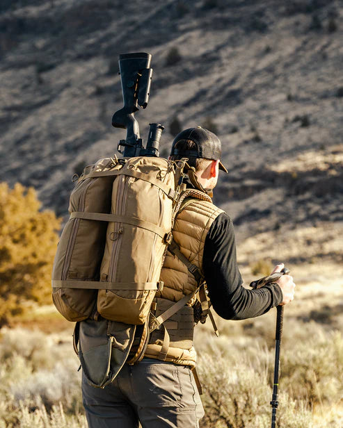 Eine Person in Outdoor-Ausrüstung steht in einer trockenen, grasbewachsenen Landschaft und blickt von der Kamera weg. Sie hält Trekkingstöcke in der Hand und trägt einen EBERLESTOCK Batwing Large - Dry Earth Rucksack, auf den ein Gewehr geschnallt ist.