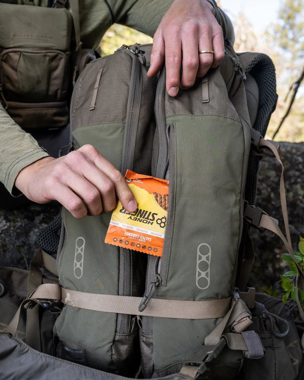 Draußen packt jemand Honey Stinger Energy Chews in das Frontfach eines grün-grauen EBERLESTOCK Eberlestock Batwing V2 Wanderrucksacks.