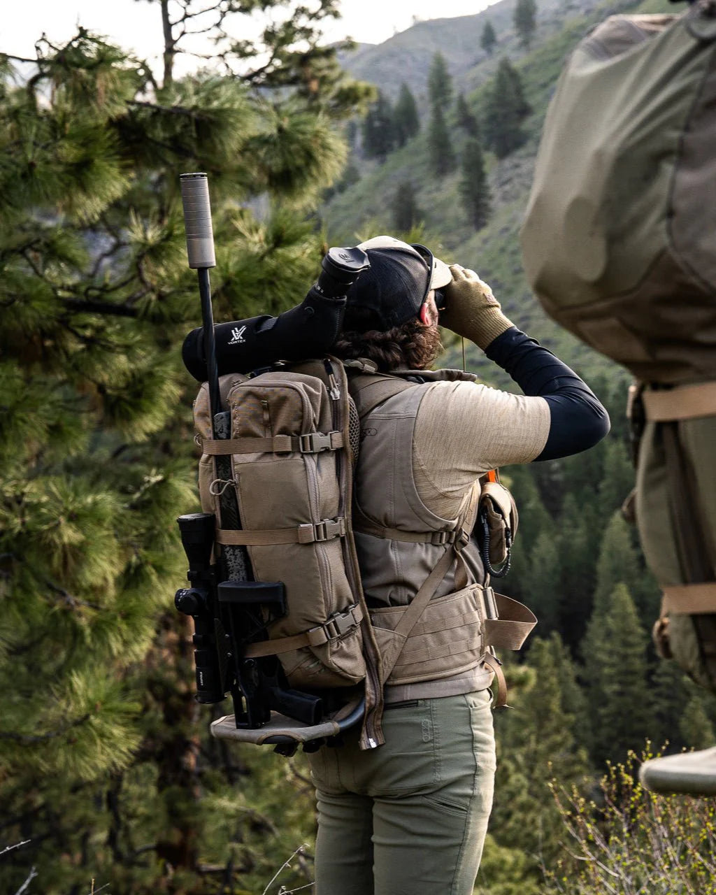 Eine Person in Outdoor-Ausrüstung und mit einem Rucksack, an dem ein EBERLESTOCK Batwing V2 befestigt ist, blickt durch ein Fernglas in eine bewaldete, bergige Gegend; ein weiterer Rucksack ist teilweise im Vordergrund zu sehen.