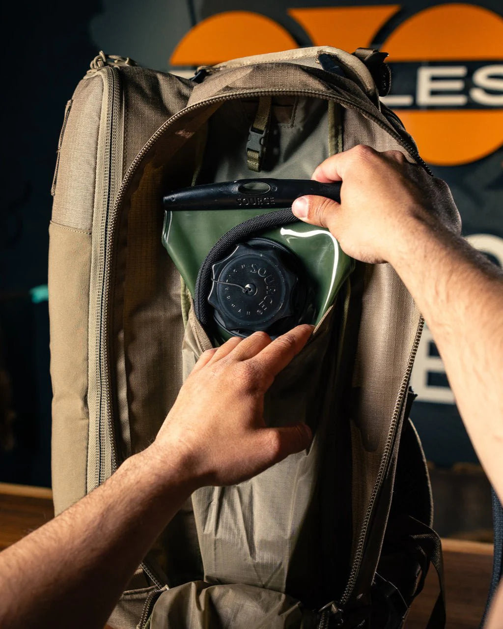 Eine Person legt ein Trinkreservoir in das Hauptfach eines EBERLESTOCK Eberlestock Batwing V2 Rucksacks, wobei sie das Reservoir mit beiden Händen hält und die Tasche teilweise geöffnet ist. Im Hintergrund erscheint ein orange-schwarzes Logo.