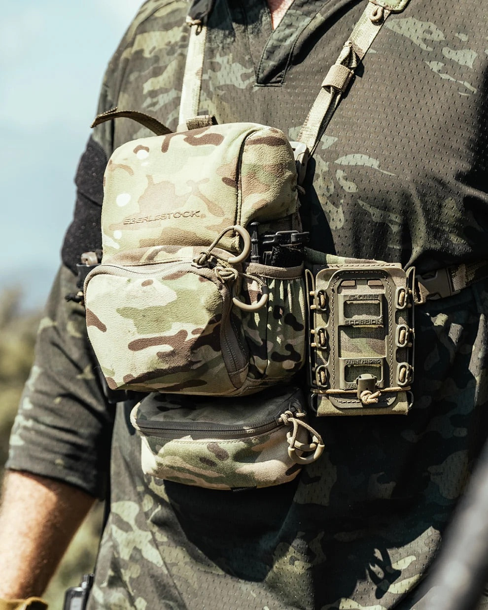 Eine Person in einer tarnfarbenen taktischen Weste mit Ausrüstung und Taschen, mit einem EBERLESTOCK Bravo Modular Bino Pack Large auf der Vorderseite. Der Hintergrund ist im Freien und leicht unscharf.