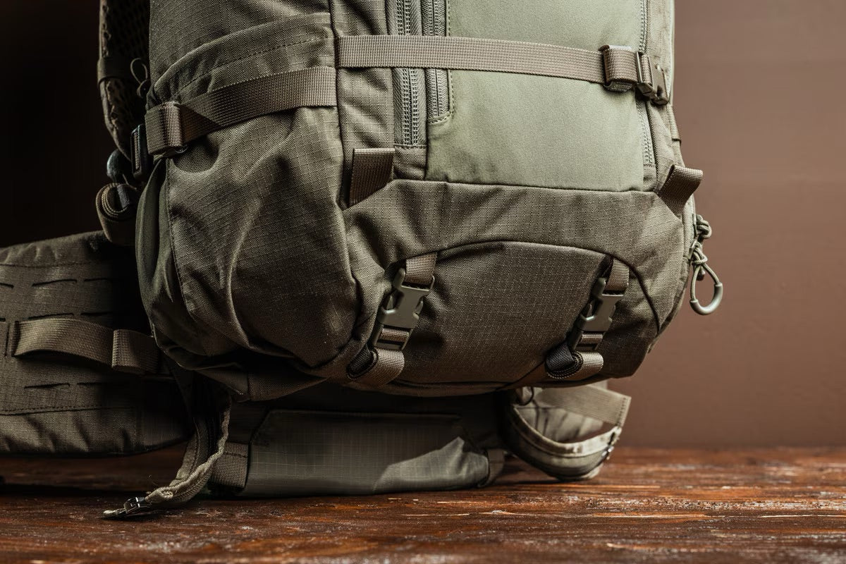 Nahaufnahme des EBERLESTOCK Brooks 3500 - Military Green Rucksacks mit Reißverschlüssen, Schnallen und Riemen auf einer Holzoberfläche und braunem Hintergrund. Dieser ultraleichte Rucksack ist robust - perfekt für Outdoor- oder Wanderabenteuer.