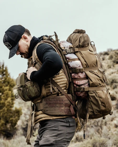 Ein Mann in Outdoor-Ausrüstung und mit Mütze wandert durch eine zerklüftete, trockene Landschaft mit spärlicher Vegetation und trägt einen mit Fleisch beladenen EBERLESTOCK Brooks 3500 - Military Green Rucksack.