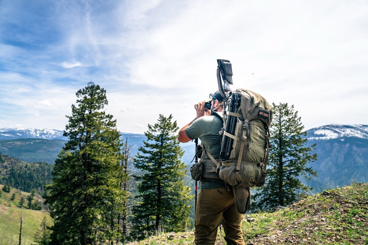 Eine Person mit einem EBERLESTOCK Brooks 7000 - Military Green Rucksack steht auf einem grasbewachsenen Hügel und betrachtet mit einem Fernglas eine Berglandschaft mit Bäumen und schneebedeckten Gipfeln unter einem teilweise bewölkten Himmel.