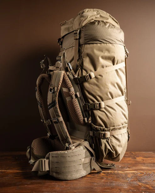Der EBERLESTOCK Brooks 7000 - Military Green Trekkingrucksack mit mehreren Gurten und gepolsterten Trägern steht aufrecht auf einer Holzunterlage, ideal für den modularen Bedarf und bereit für jedes Abenteuer auf braunem Grund.