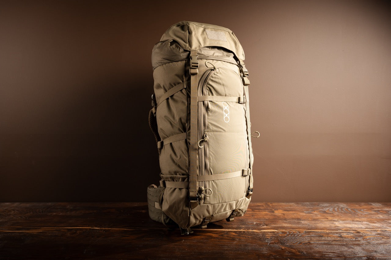 Der EBERLESTOCK Brooks 7000 - Military Green ist ein großer modularer Rucksack mit mehreren Riemen und Schnallen, der aufrecht auf einer hölzernen Oberfläche vor einem schlichten braunen Hintergrund steht.