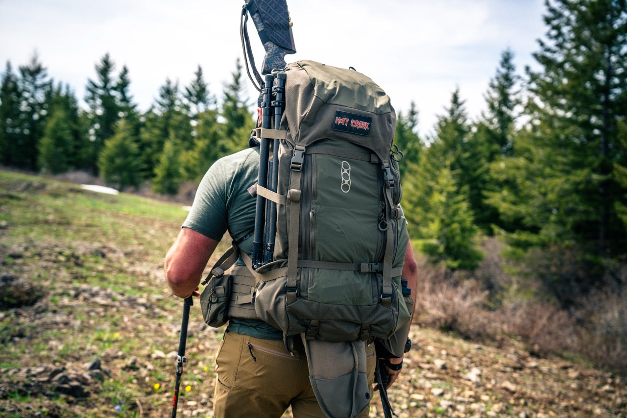 Eine Person mit einem EBERLESTOCK Brooks 7000 - Military Green Rucksack und einem Gewehr, die auf einem felsigen Pfad inmitten von Bäumen und Grün wandern - bereit für ihr nächstes Trekking-Abenteuer.