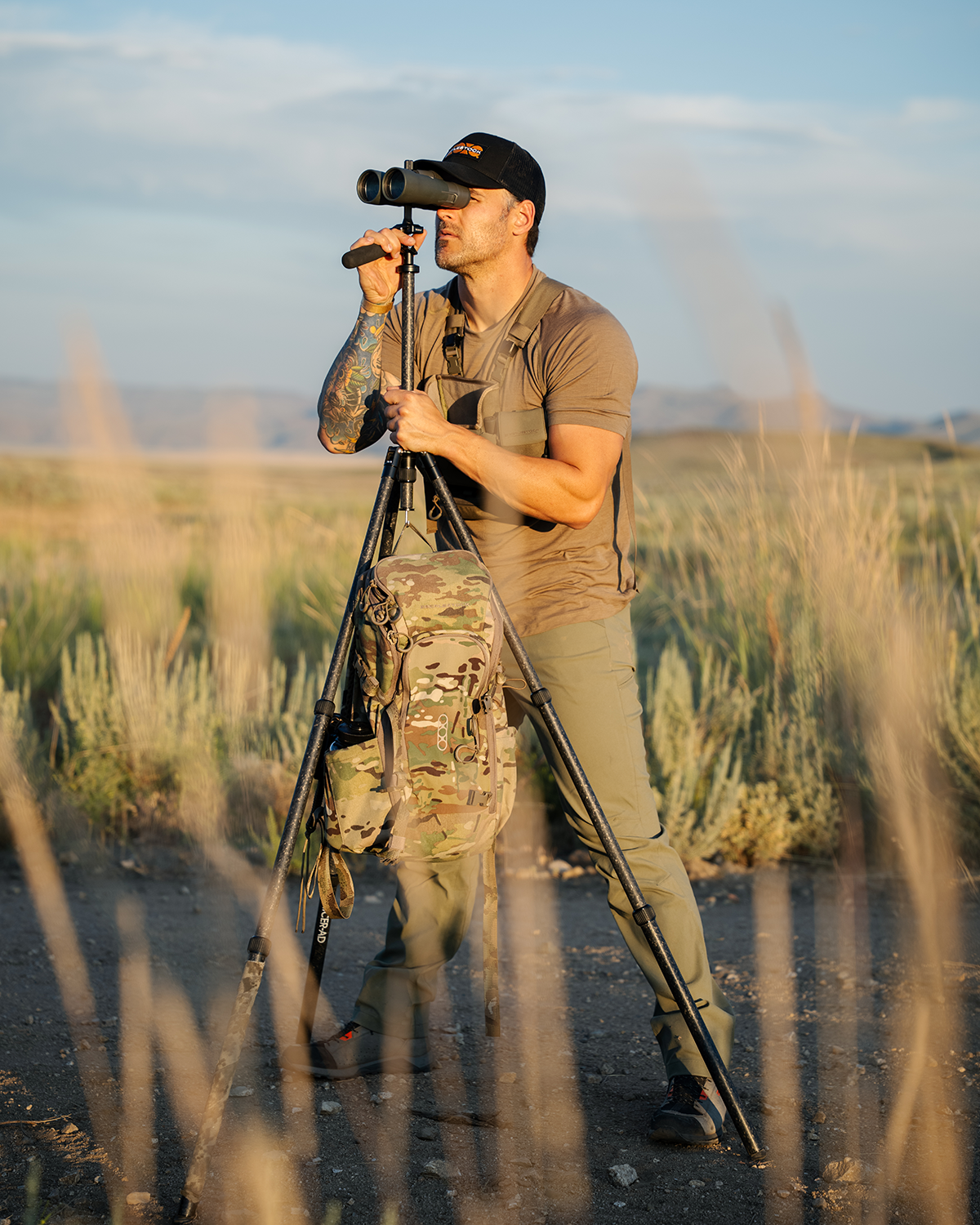 Ein Mann in Outdoor-Ausrüstung blickt durch ein auf einem Stativ montiertes Fernglas in ein sonniges Feld. Er trägt eine Mütze und das EBERLESTOCK Brute Scout, mit einer Tarntasche auf dem Stativ. Im Hintergrund erheben sich Hügel.