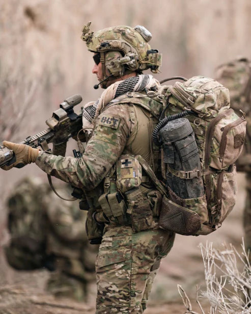 Ein Soldat in tarnfarbener Uniform und taktischer Ausrüstung, mit Helm und Handschuhen, trägt einen EBERLESTOCK FAC Track - Coyote Brown Rucksack mit MOLLE-System, während er im Freien spazieren geht und sich in das raue natürliche Gelände einfügt.